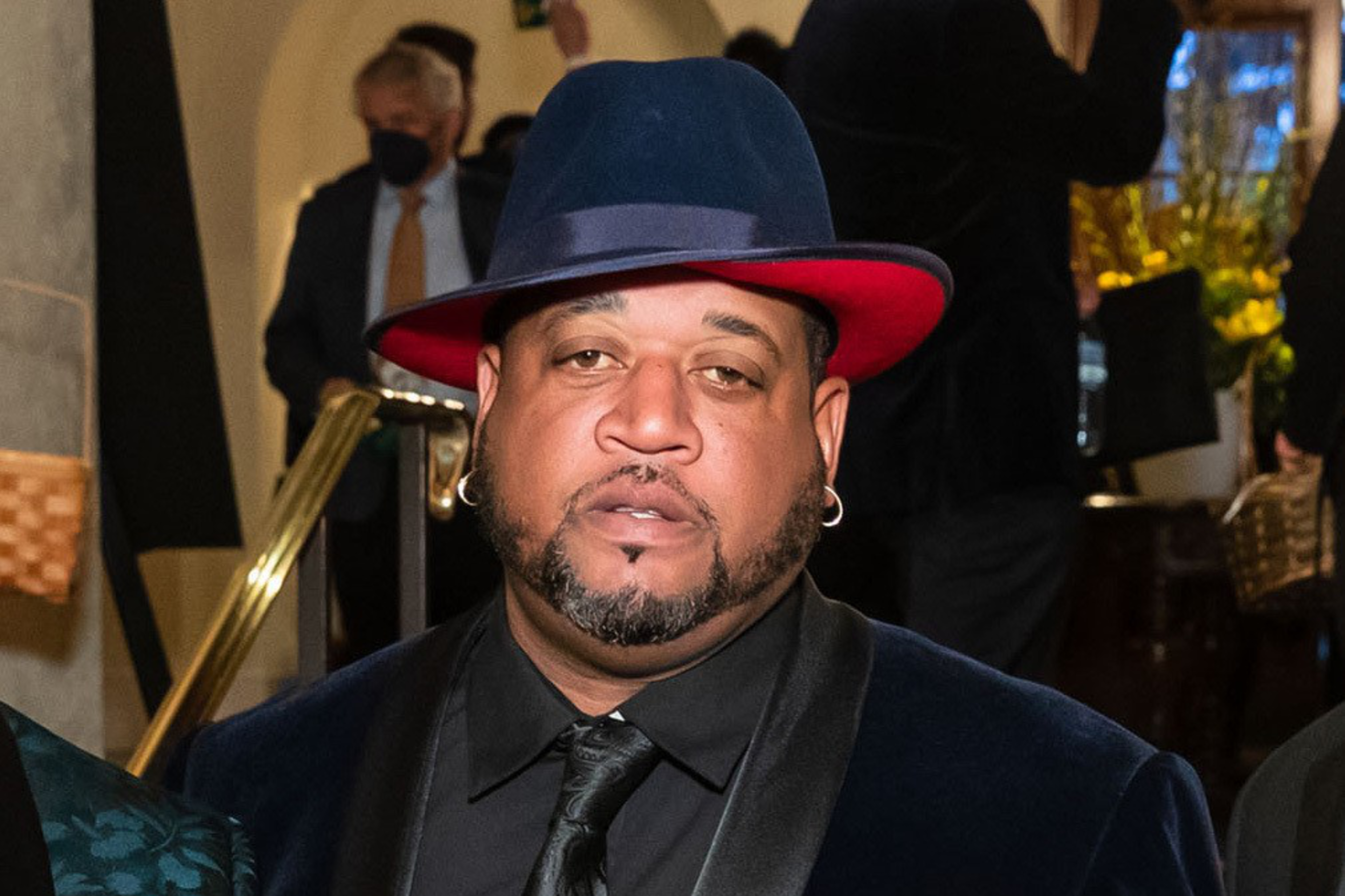 A man in a blue hat with a red brim, wearing earrings and a dark suit, is in an indoor setting with other people nearby.