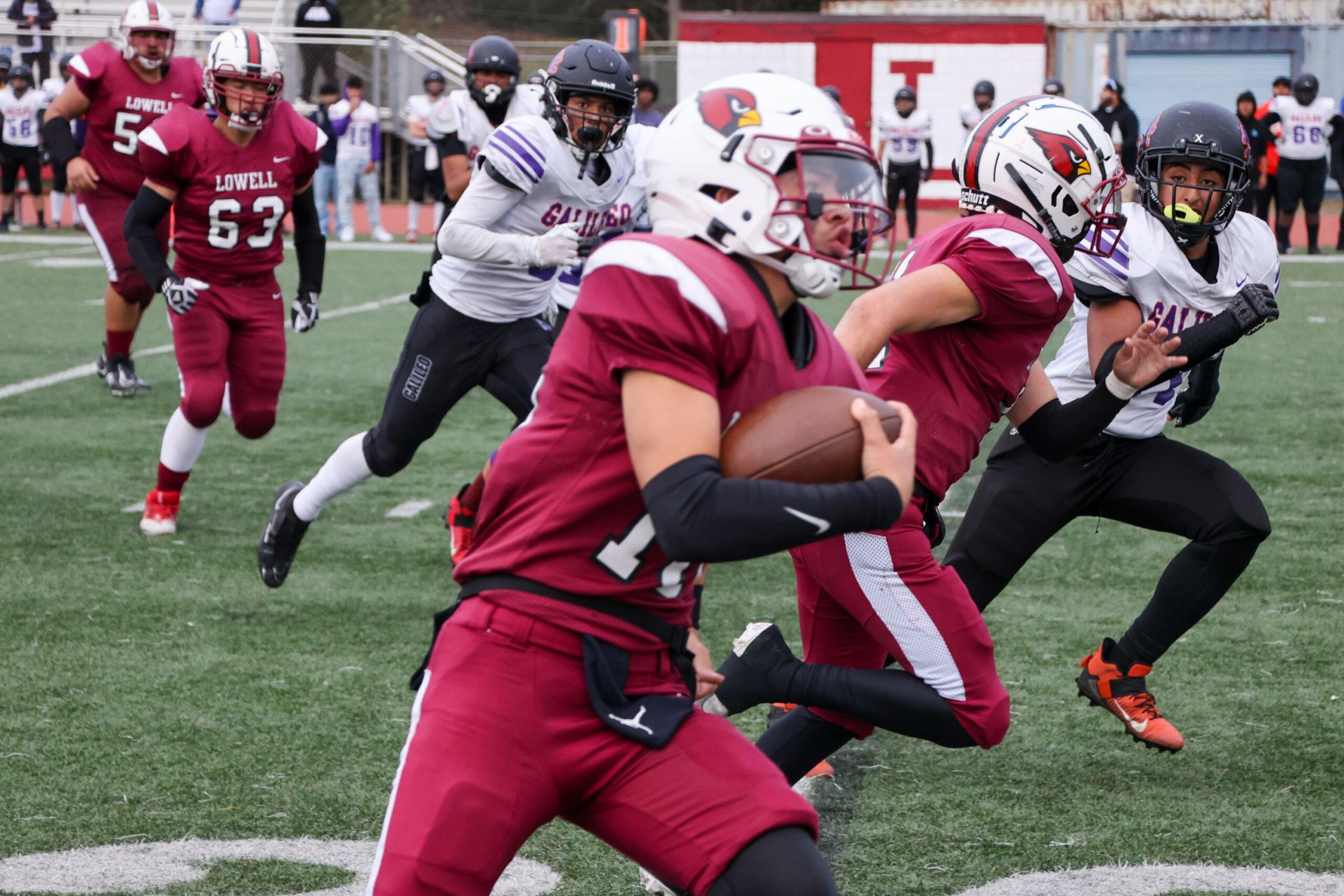 A football player in maroon runs with the ball as opponents in white and black attempt to tackle him on a green field.
