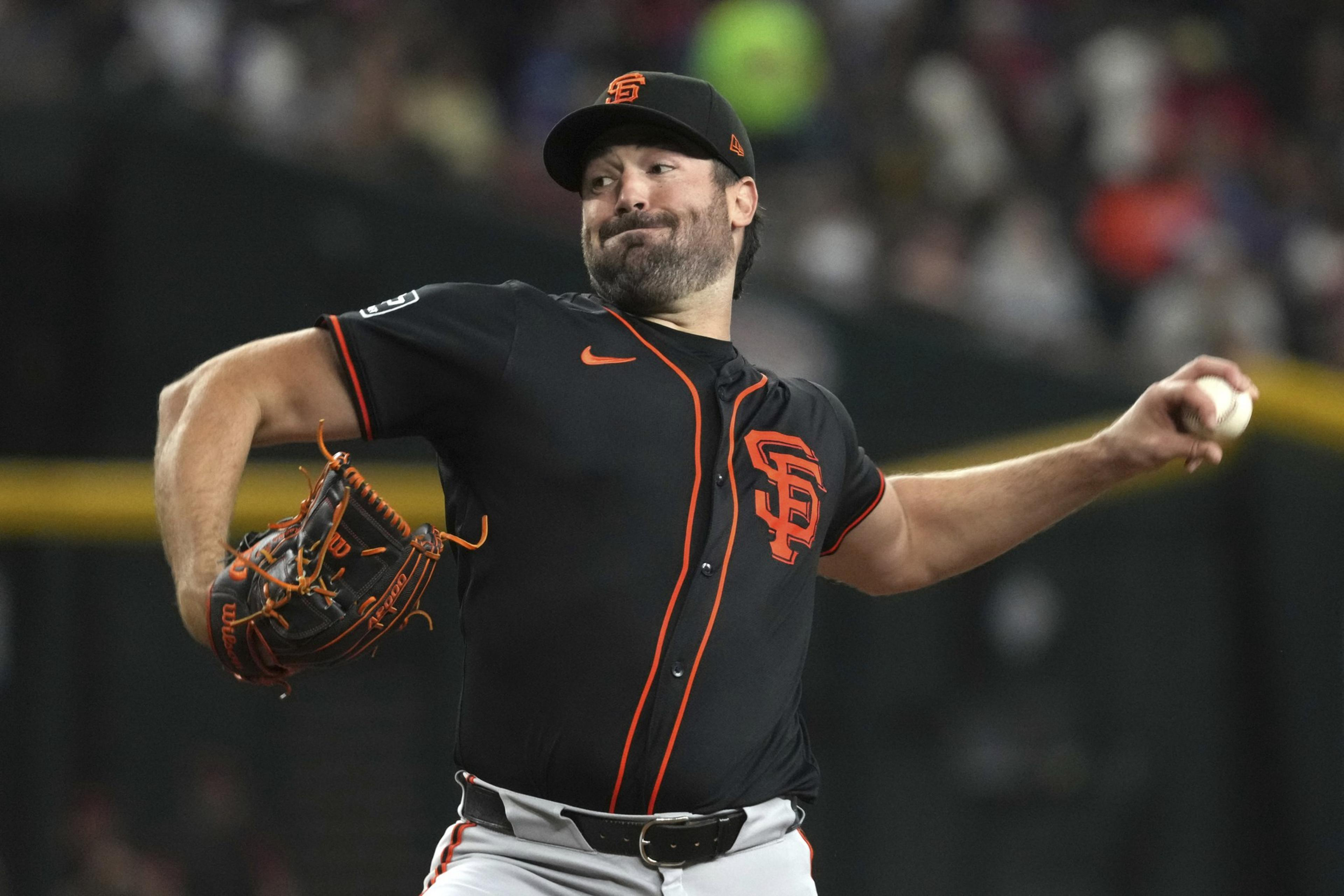 A baseball player is pitching in a game, wearing a black and orange uniform and cap with "SF" lettering. He's mid-throw, with a focused expression.