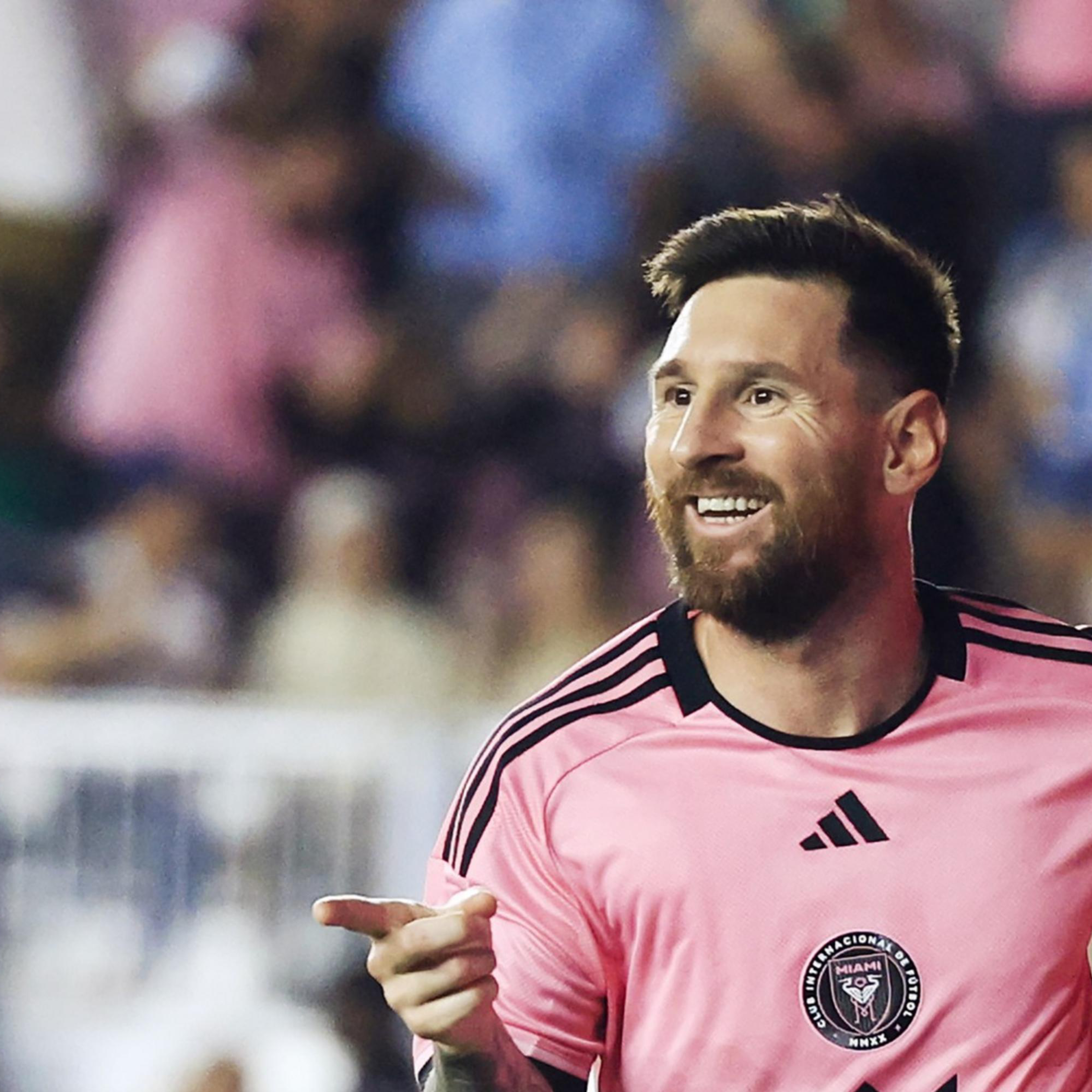 A soccer player in a pink jersey is smiling and pointing, with an enthusiastic crowd in the background.