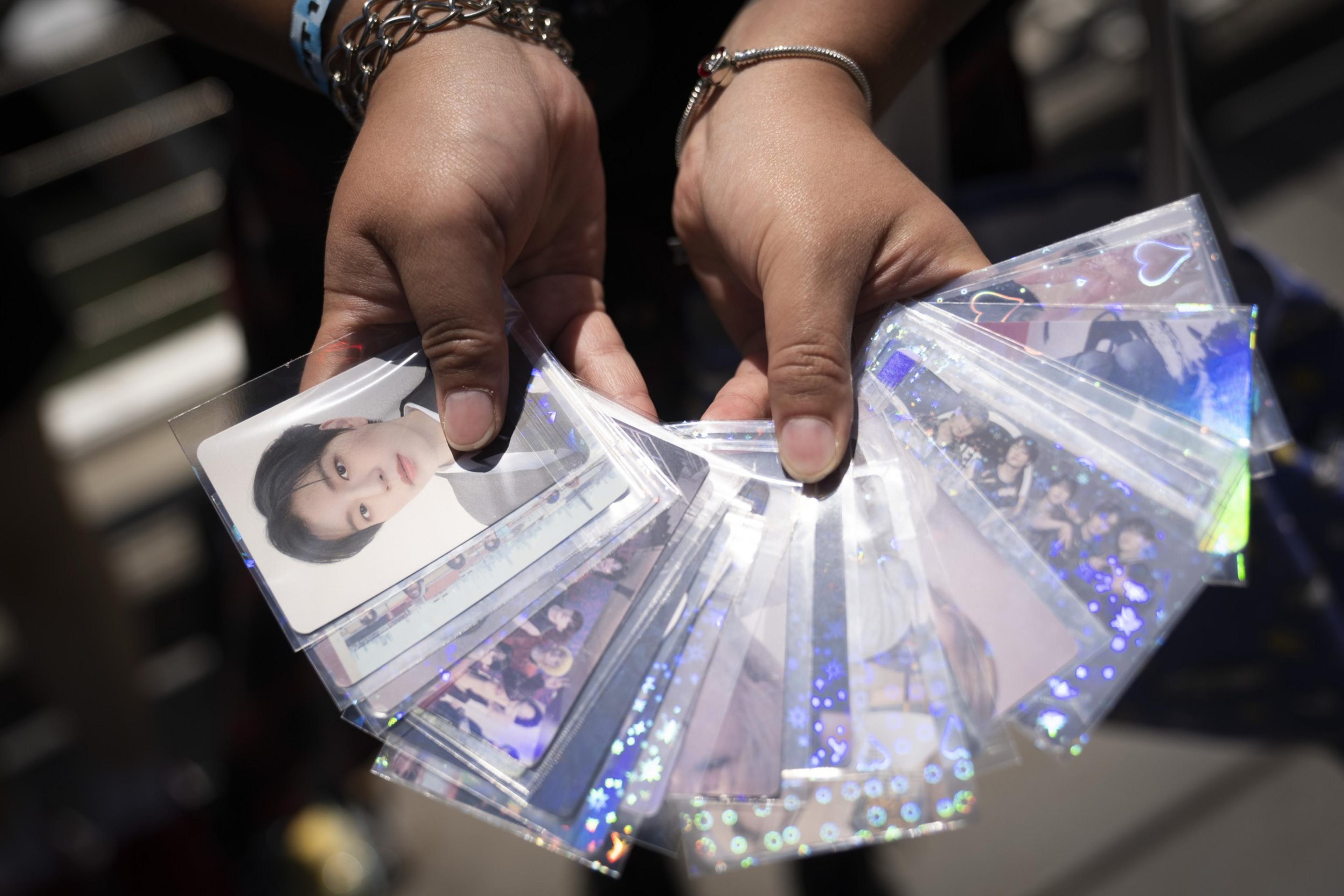 Two hands hold a fan of K-pop photo cards featuring people in sparkling sleeves. A wristband and bracelet are visible on the left wrist.