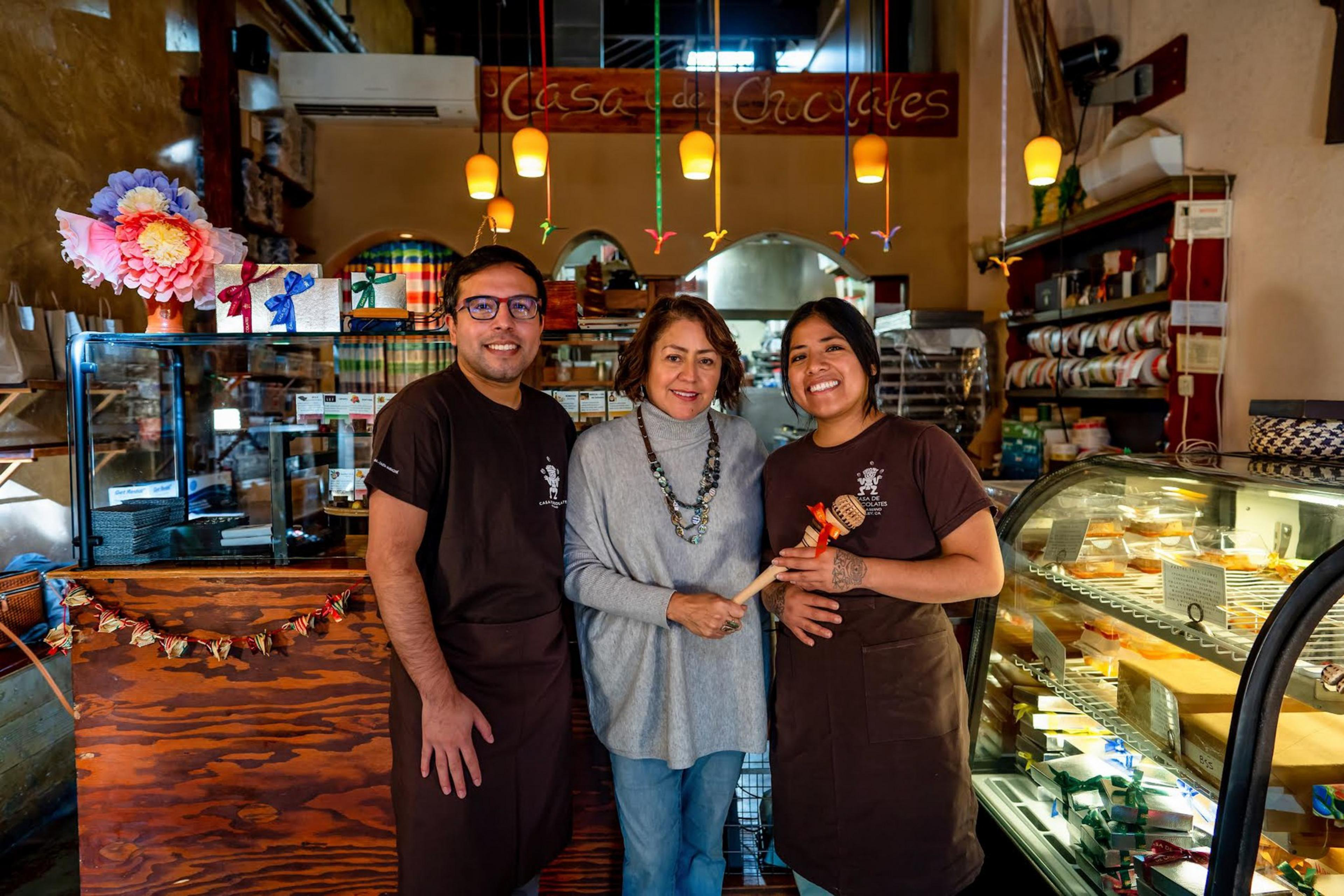 Three people smile inside a rustic chocolate shop; two are wearing matching brown aprons and the one in the middle wears a grey sweater and jeans.