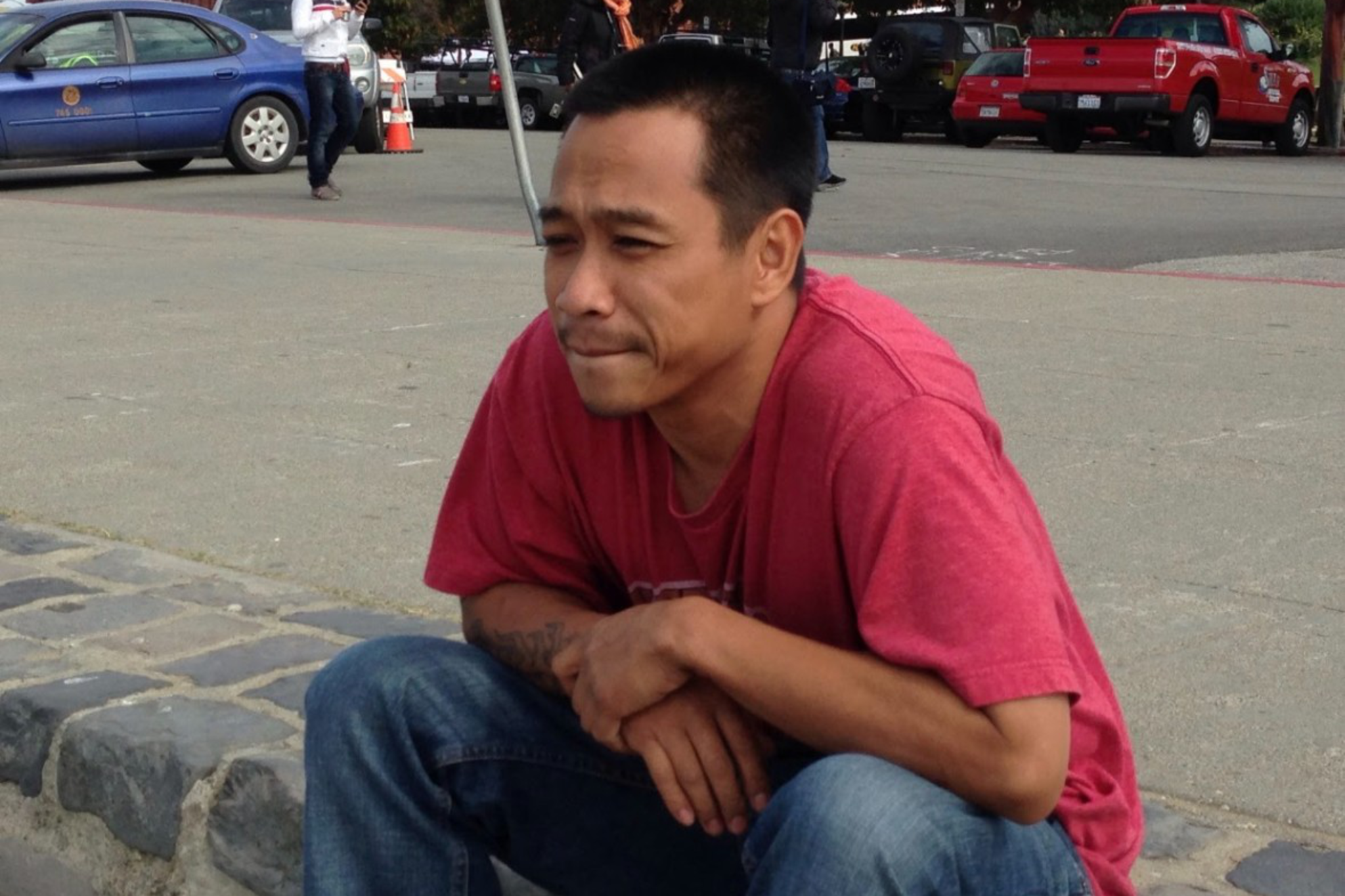 A person in a red T-shirt is crouching on a pavement near parked cars and people. They appear deep in thought, with a street and building in the background.