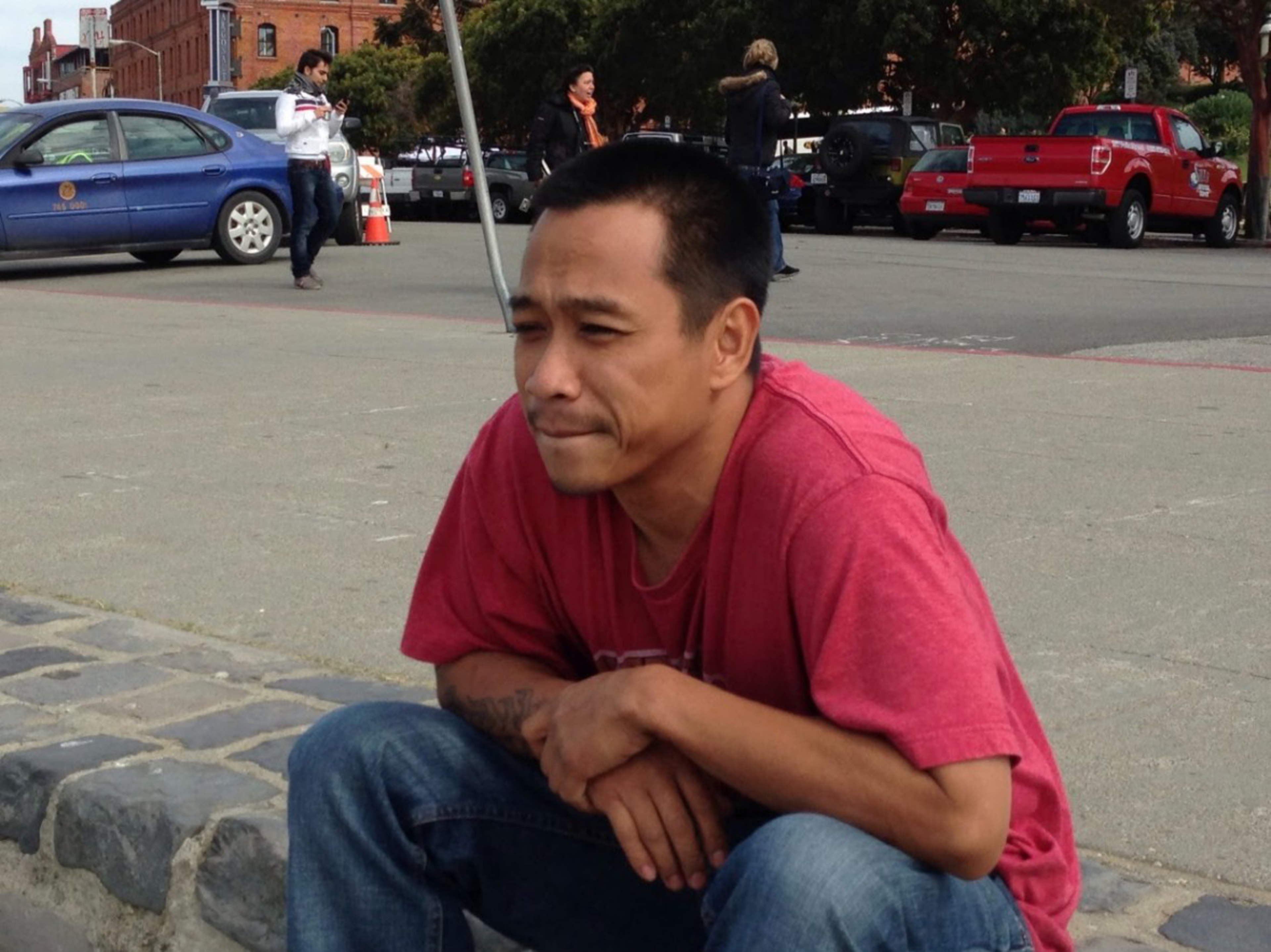 A person in a red T-shirt is crouching on a pavement near parked cars and people. They appear deep in thought, with a street and building in the background.
