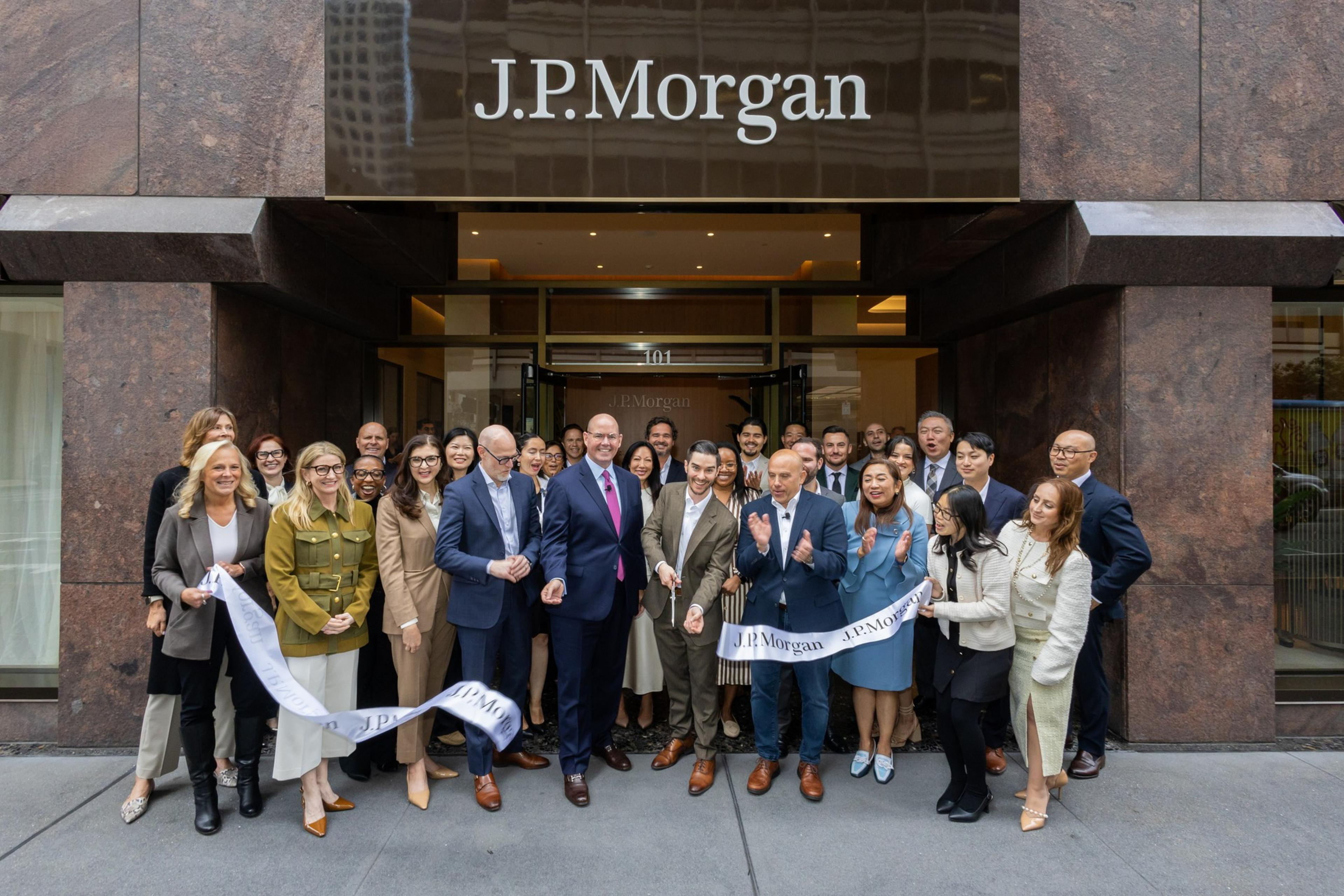 A group of professionally dressed people are gathered in front of a J.P. Morgan building for a ribbon-cutting ceremony, holding a ribbon and smiling.