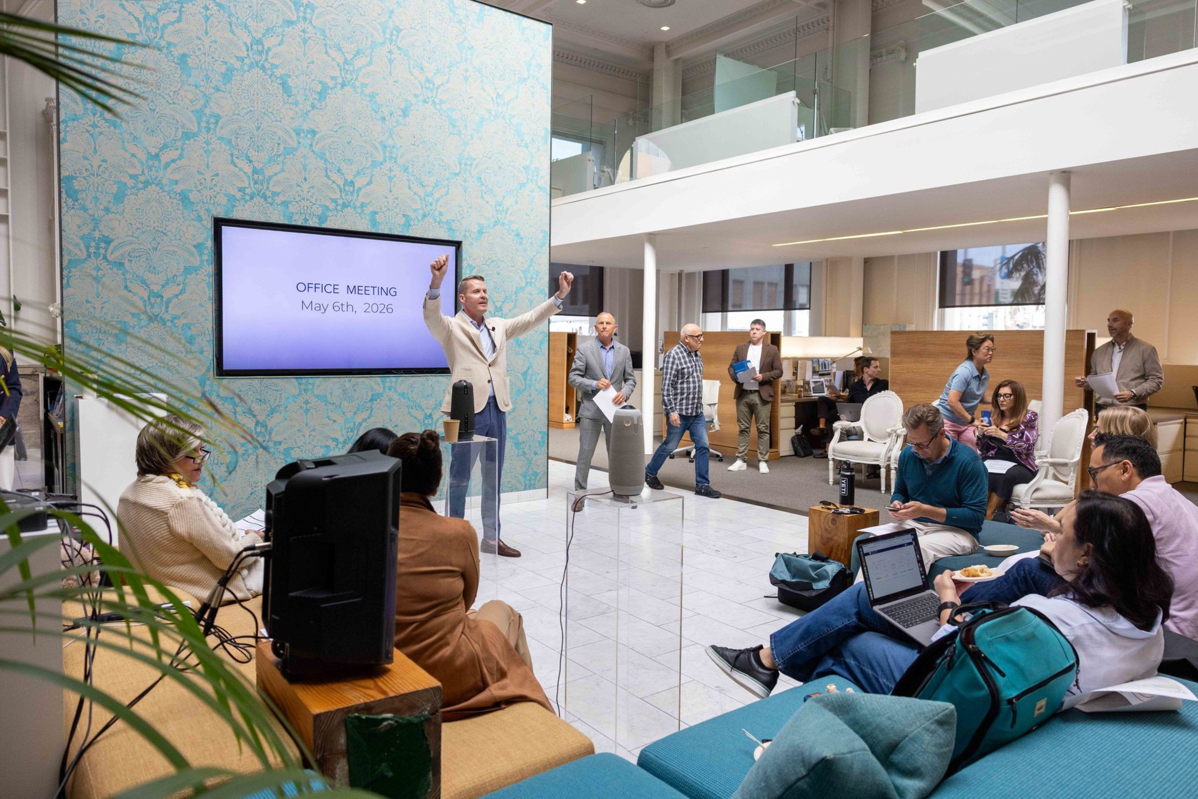 A man in a suit gestures during an office meeting with a screen displaying “May 6th, 2026,” while colleagues sit, listen, and work on laptops.