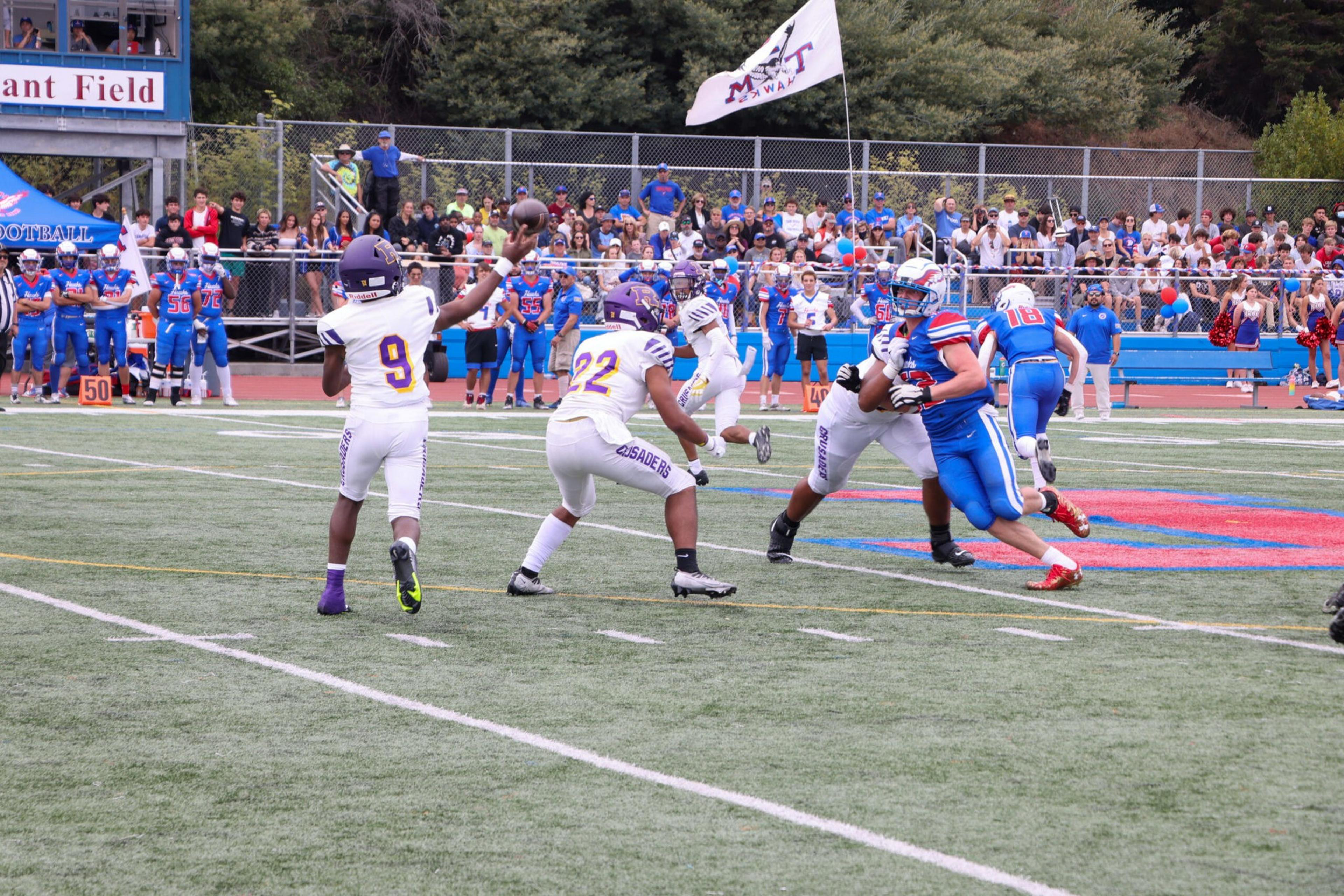 A football quarterback wearing number 9 in white throws a pass as number 22 blocks a blue team player, with a crowd and players on the sidelines watching.