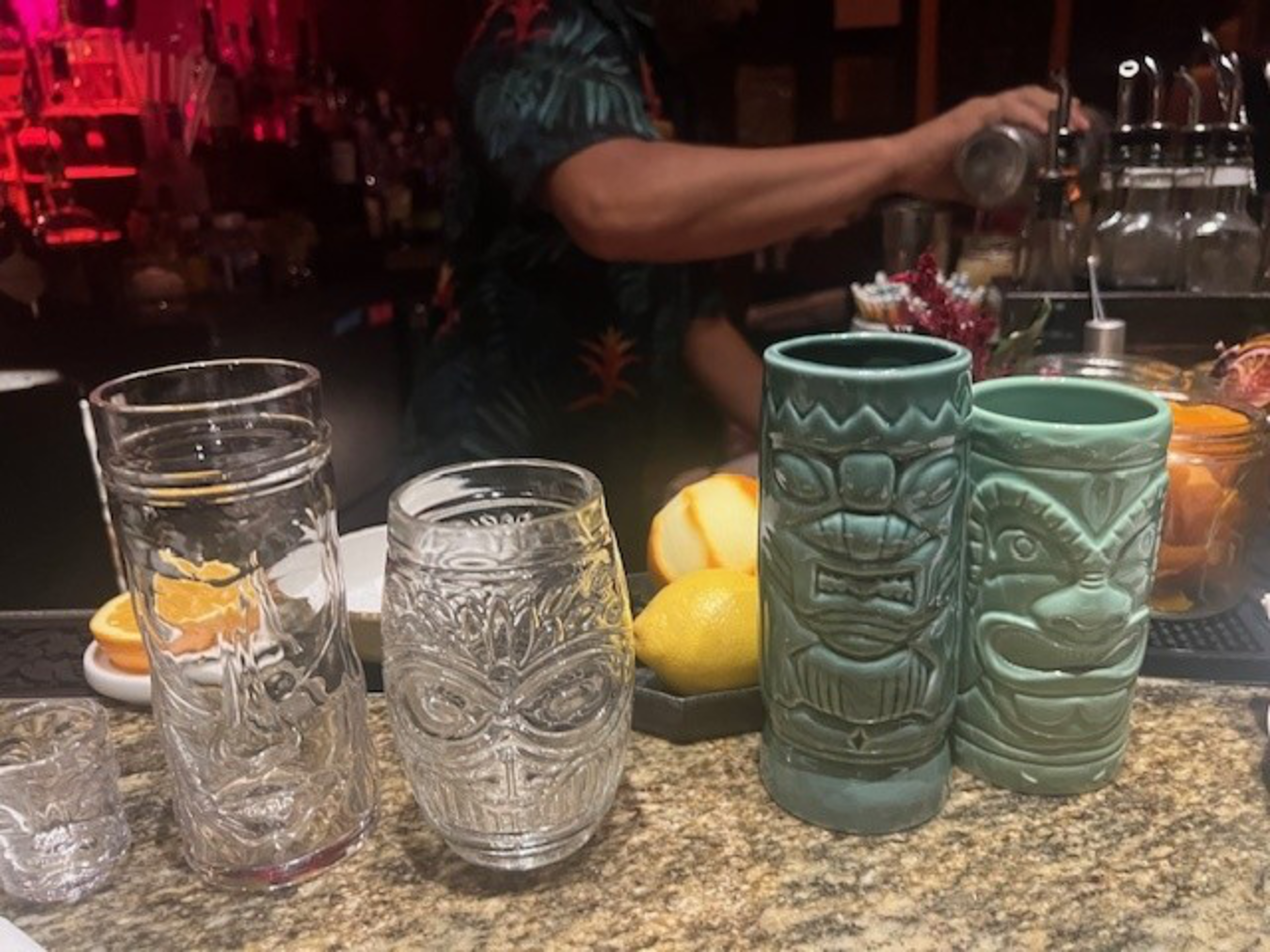 Two clear glass tiki mugs stand next to two ceramic mugs on a bar top.