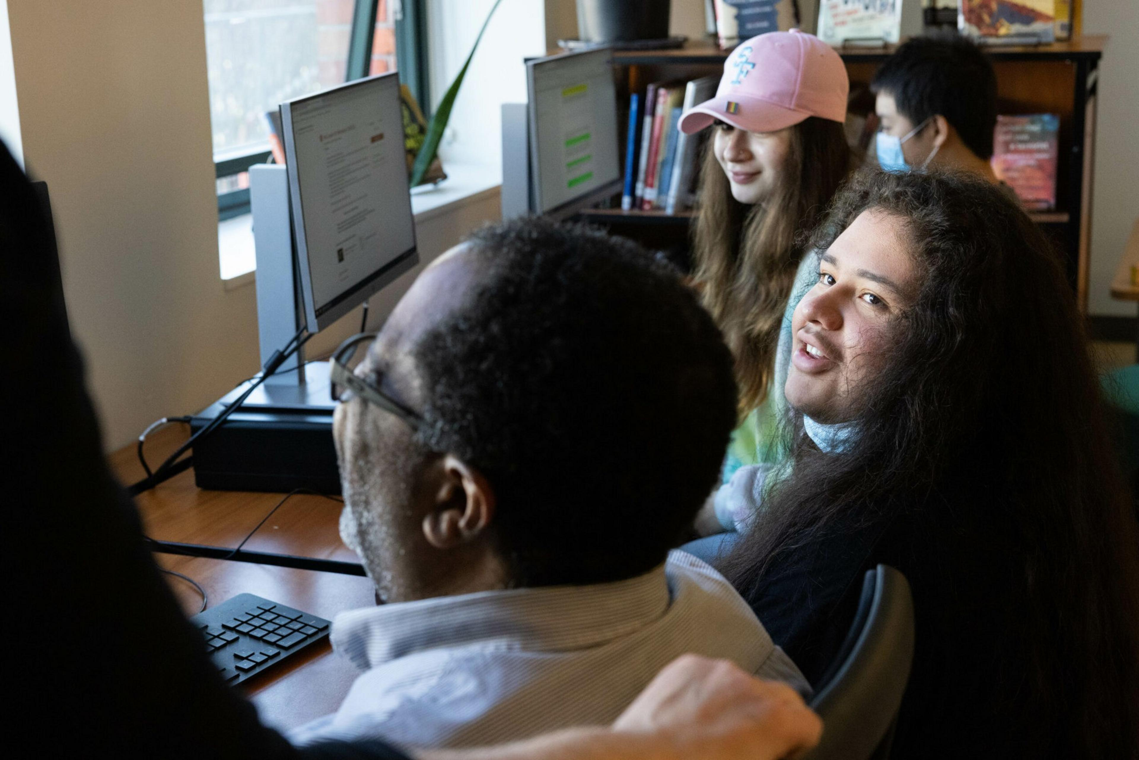 Four people sit at a desk with computer monitors, one man looks up while others focus on the screens, a bookshelf and window are visible behind them.