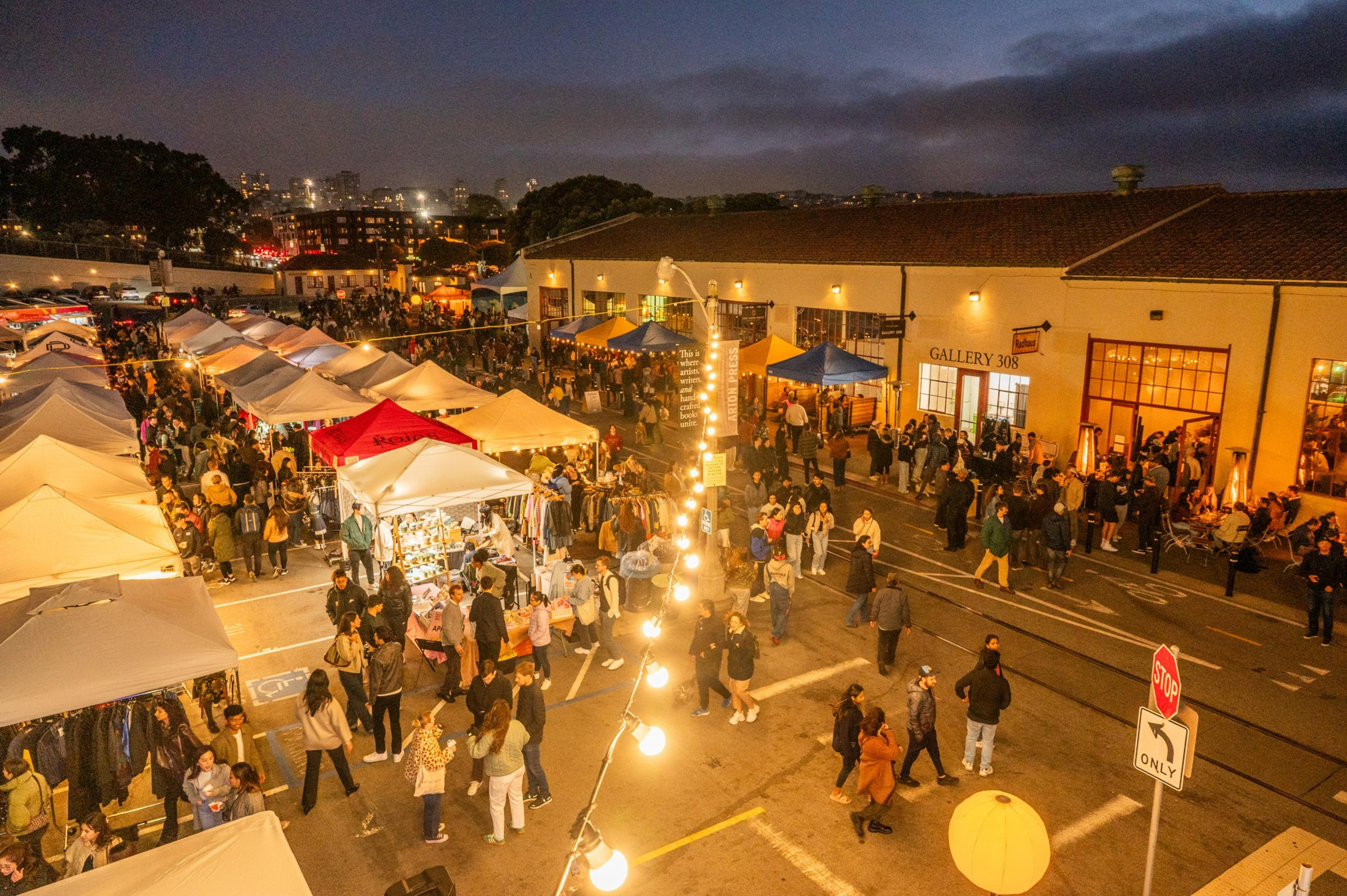 A bustling night market with rows of illuminated tents, vendors selling goods, and a lively crowd. Warm light strings add ambiance, with a city backdrop.