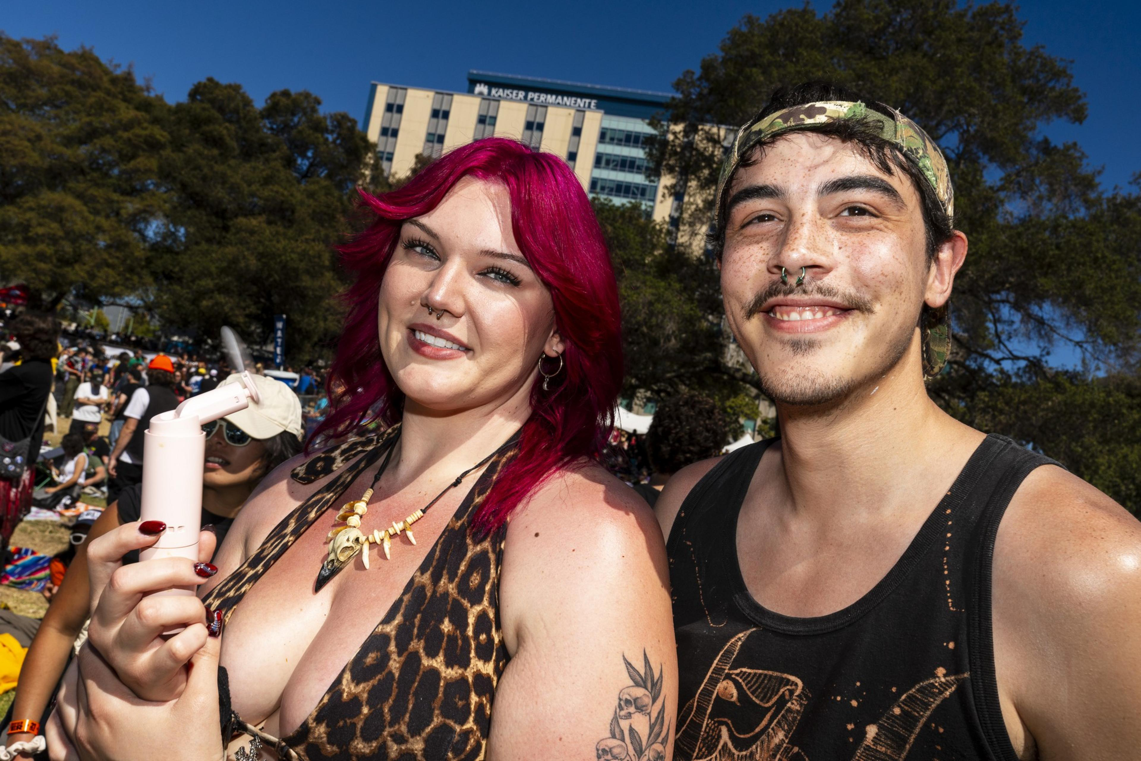 A woman with pink hair and a leopard print outfit holds a device, smiling beside a man in a cap. They're outdoors, with trees and a building behind them.