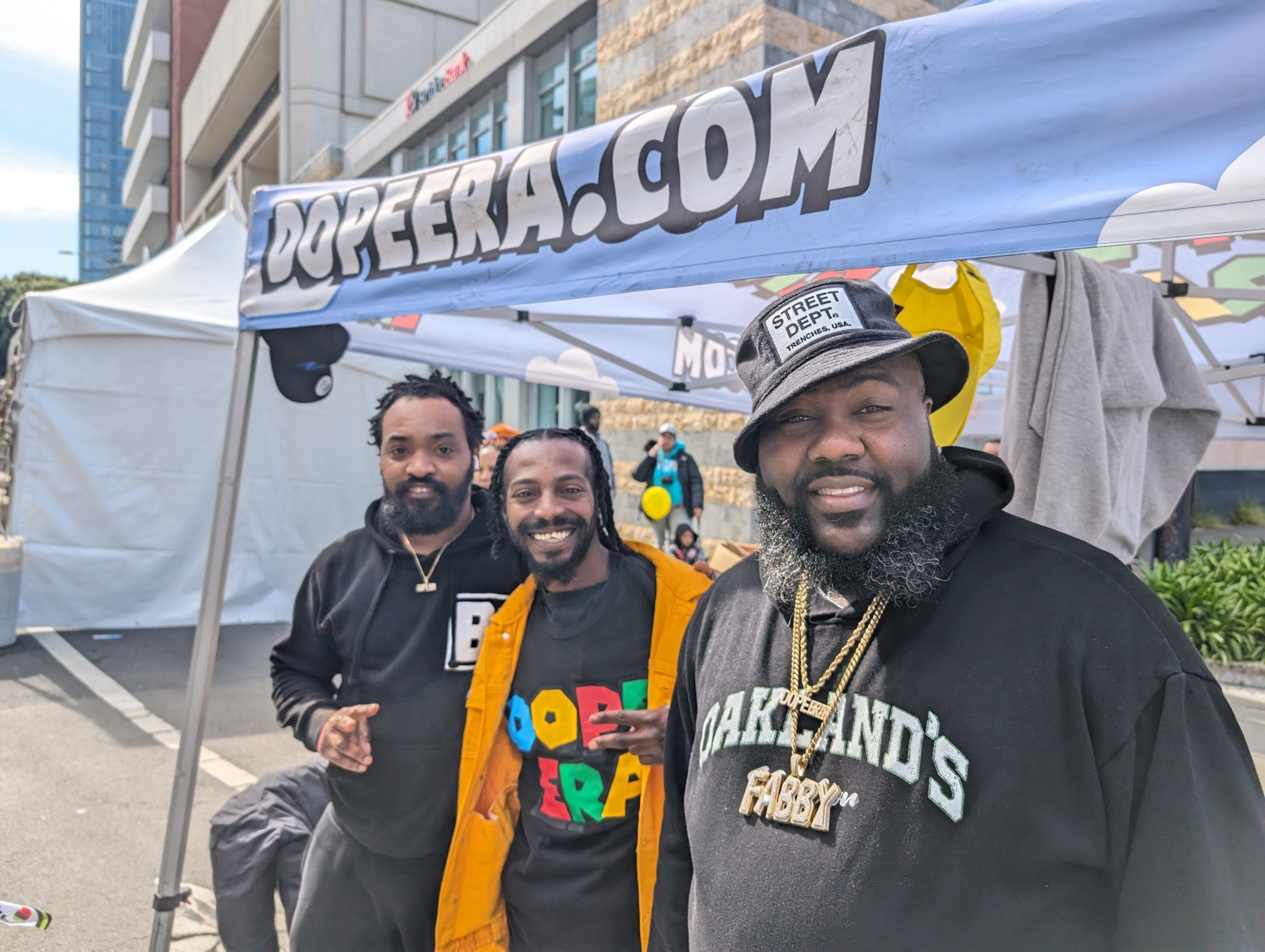 Three men in stylish casual clothing stand under an outdoor festival booth.