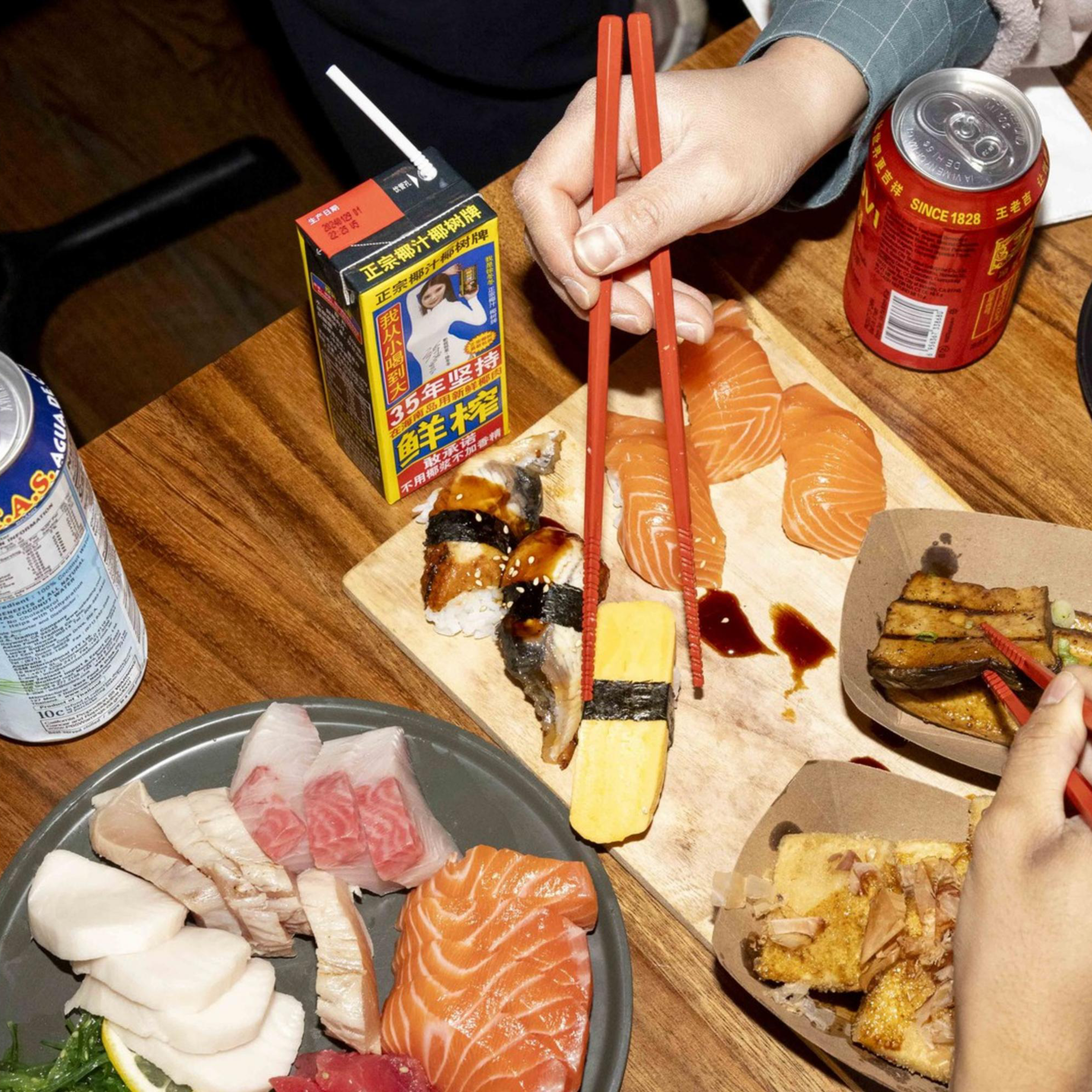 A table is filled with various Japanese dishes, including sushi, sashimi, rolls, and a bowl of bright yellow corn. Several hands holding chopsticks reach for the food.