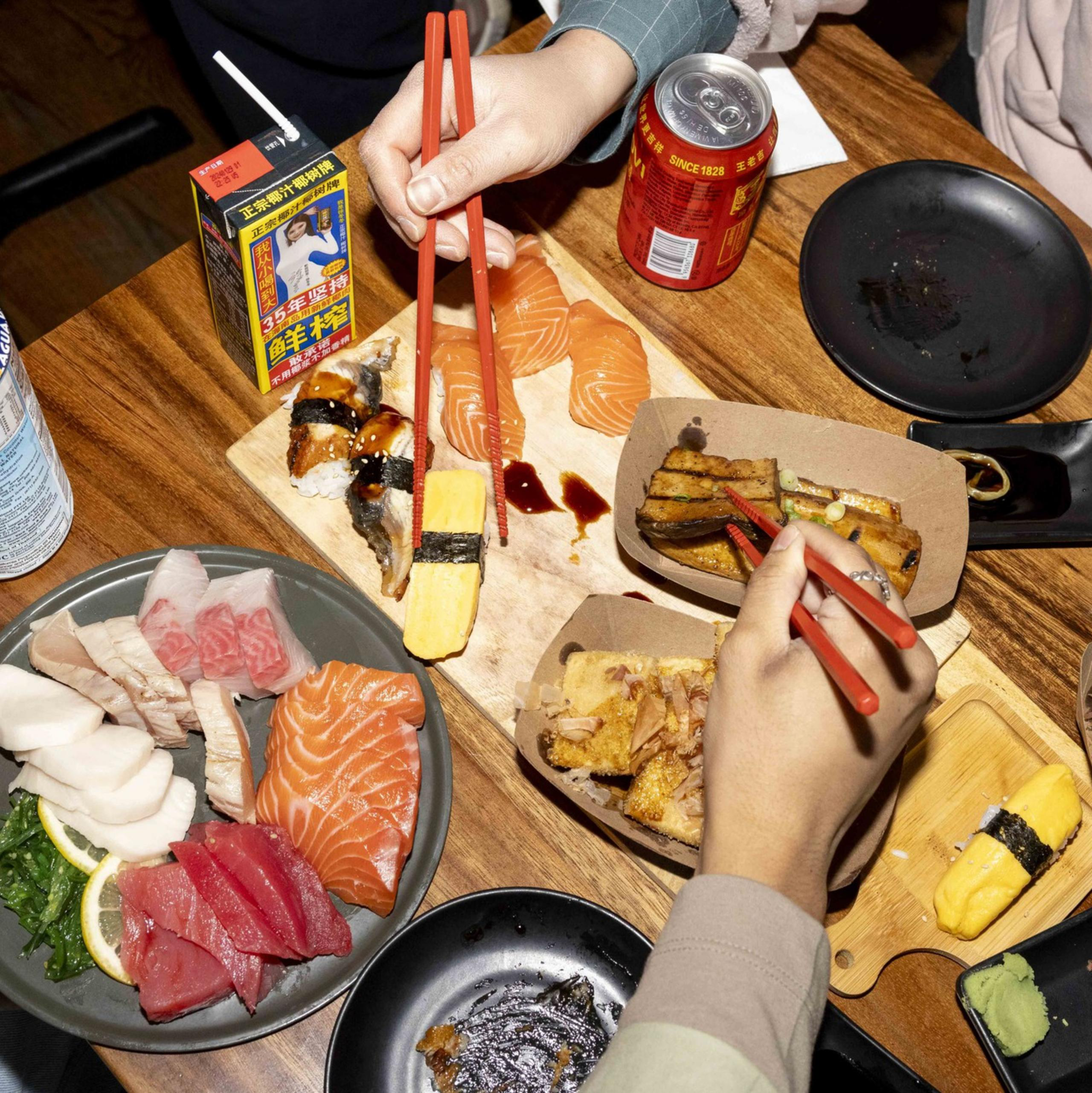 A table is filled with various Japanese dishes, including sushi, sashimi, rolls, and a bowl of bright yellow corn. Several hands holding chopsticks reach for the food.