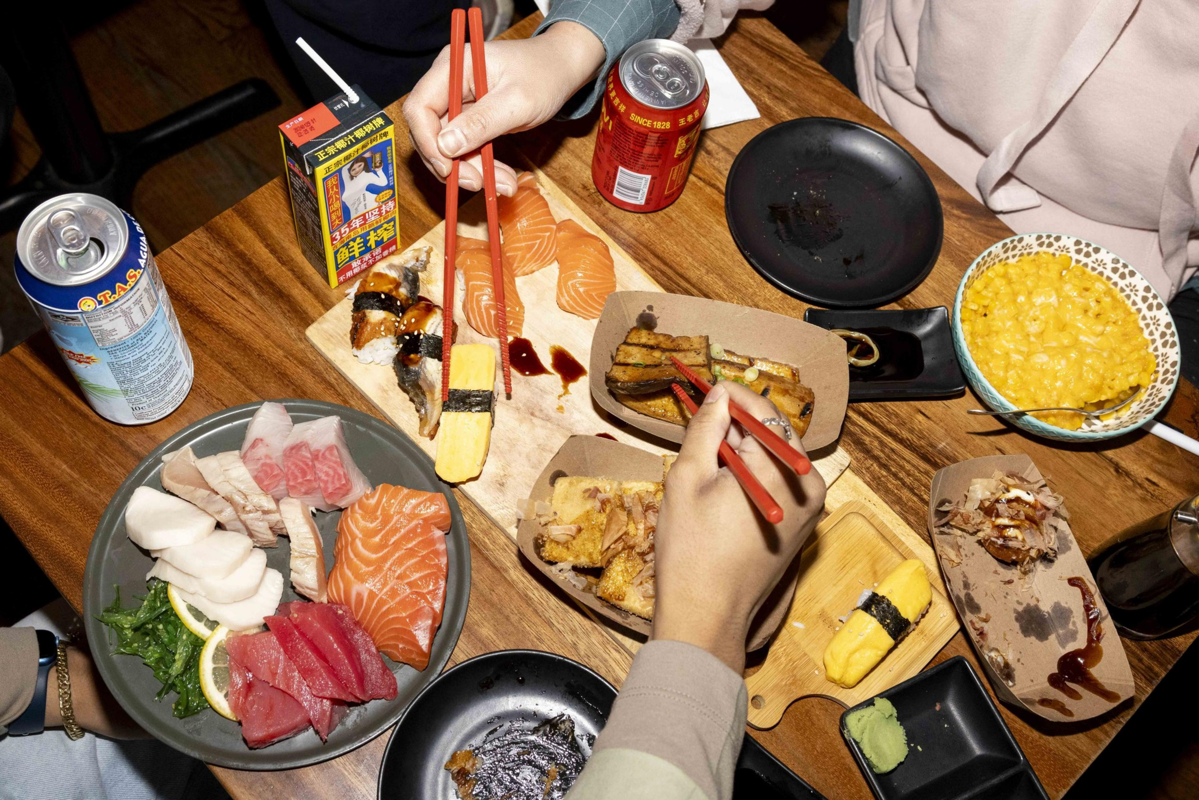 A table is filled with various Japanese dishes, including sushi, sashimi, rolls, and a bowl of bright yellow corn. Several hands holding chopsticks reach for the food.