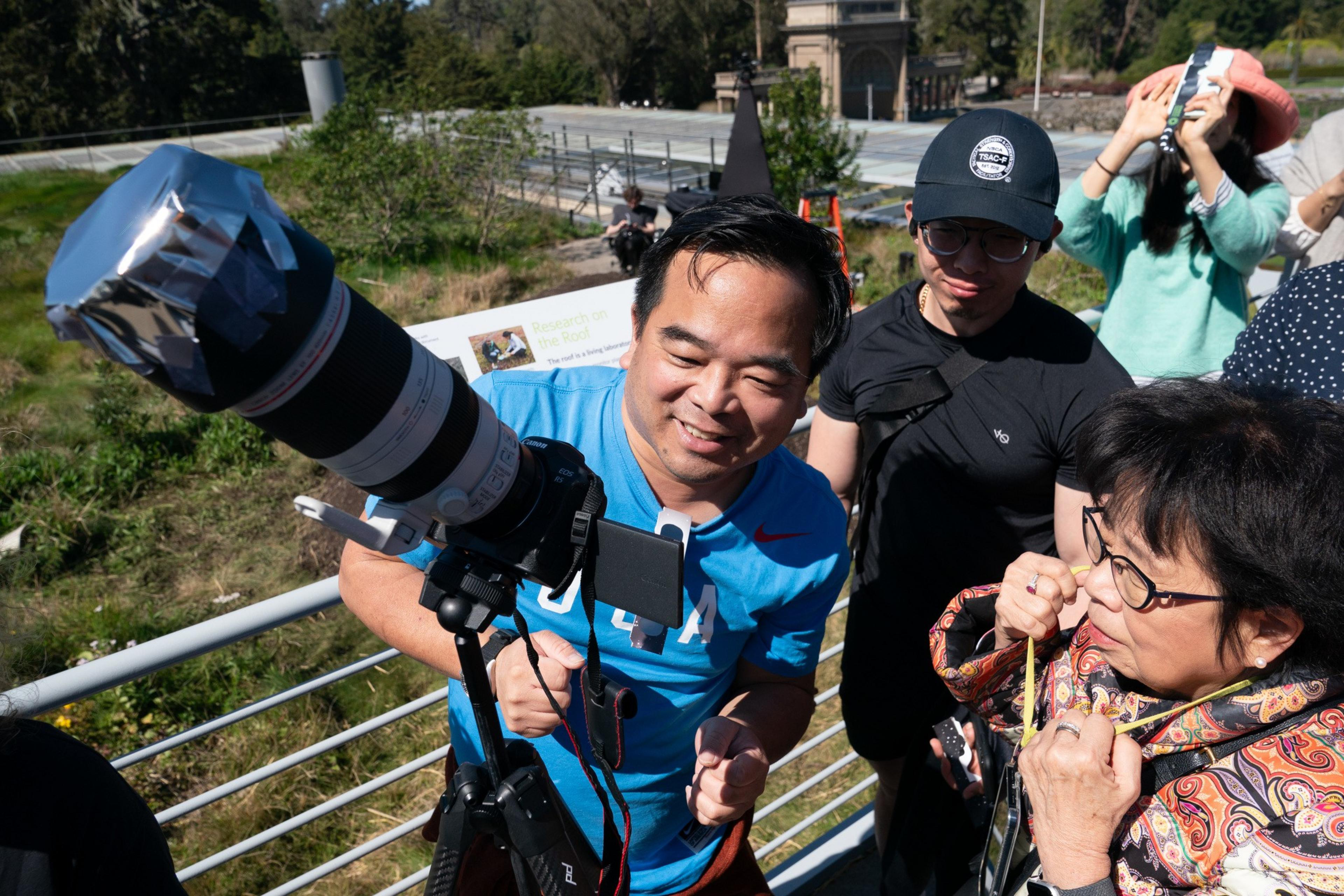 A man smiles while operating a large camera on a tripod, with interested onlookers behind him in daylight.