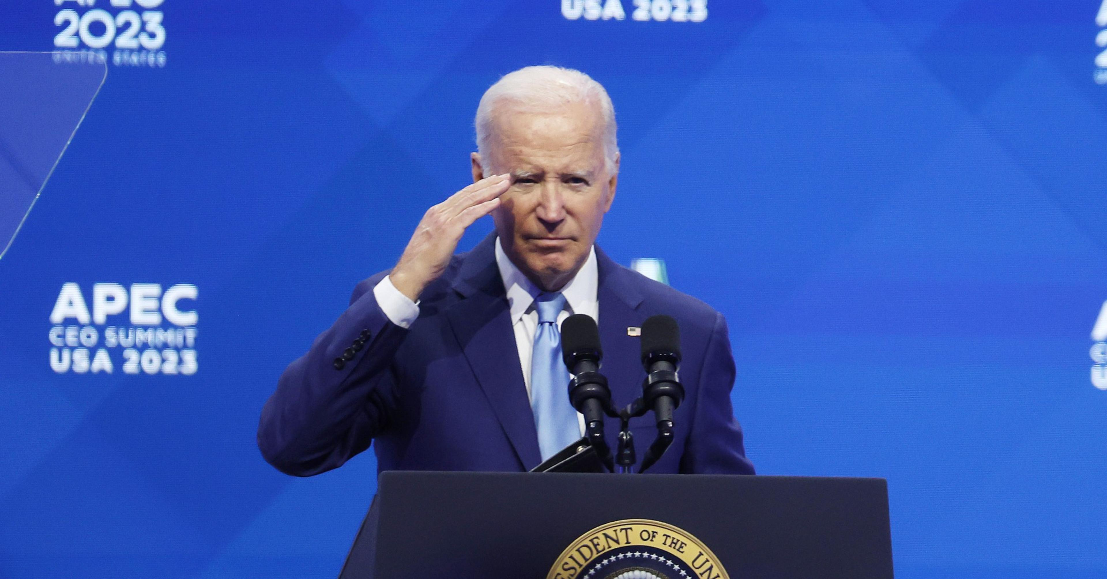U.S. President Joe Biden stands behind a lectern saluting with a blue backdrop in the background that reads: APEC CEO SUMMIT USA 2023