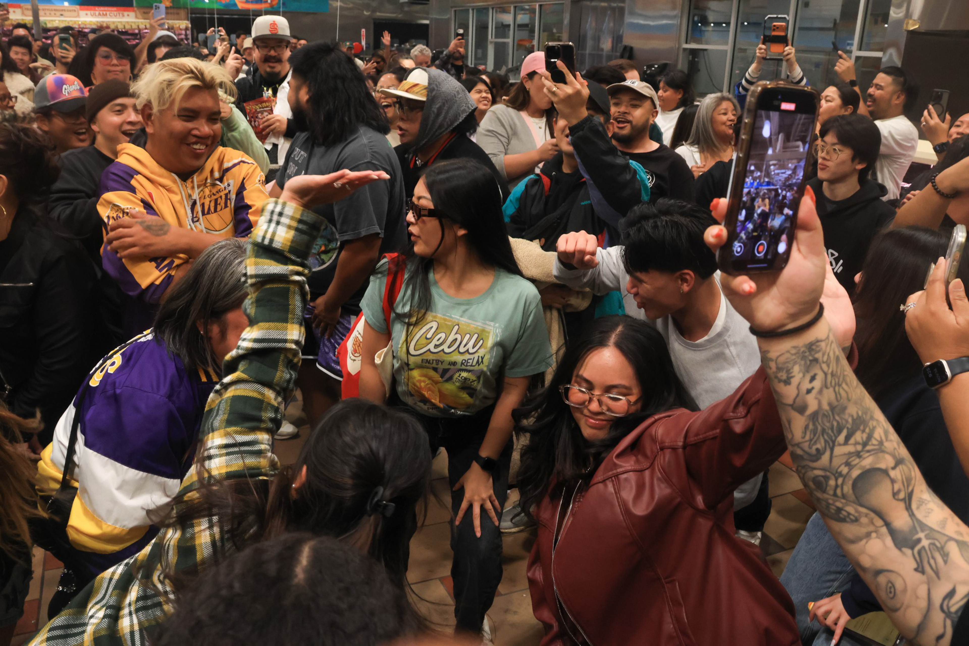 A lively crowd of diverse people dance closely together, some smiling, others recording the moment on phones in an indoor, festive atmosphere.