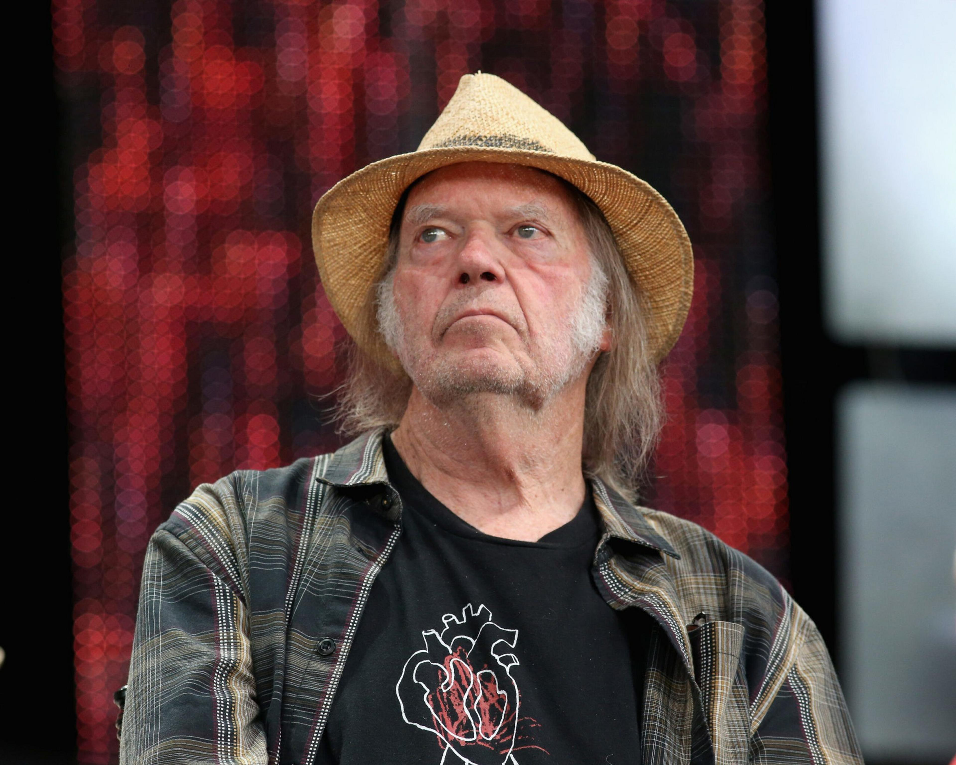An older man with long gray hair and mutton chops wears a straw hat, a plaid shirt, and a black t-shirt with a heart and flames design.