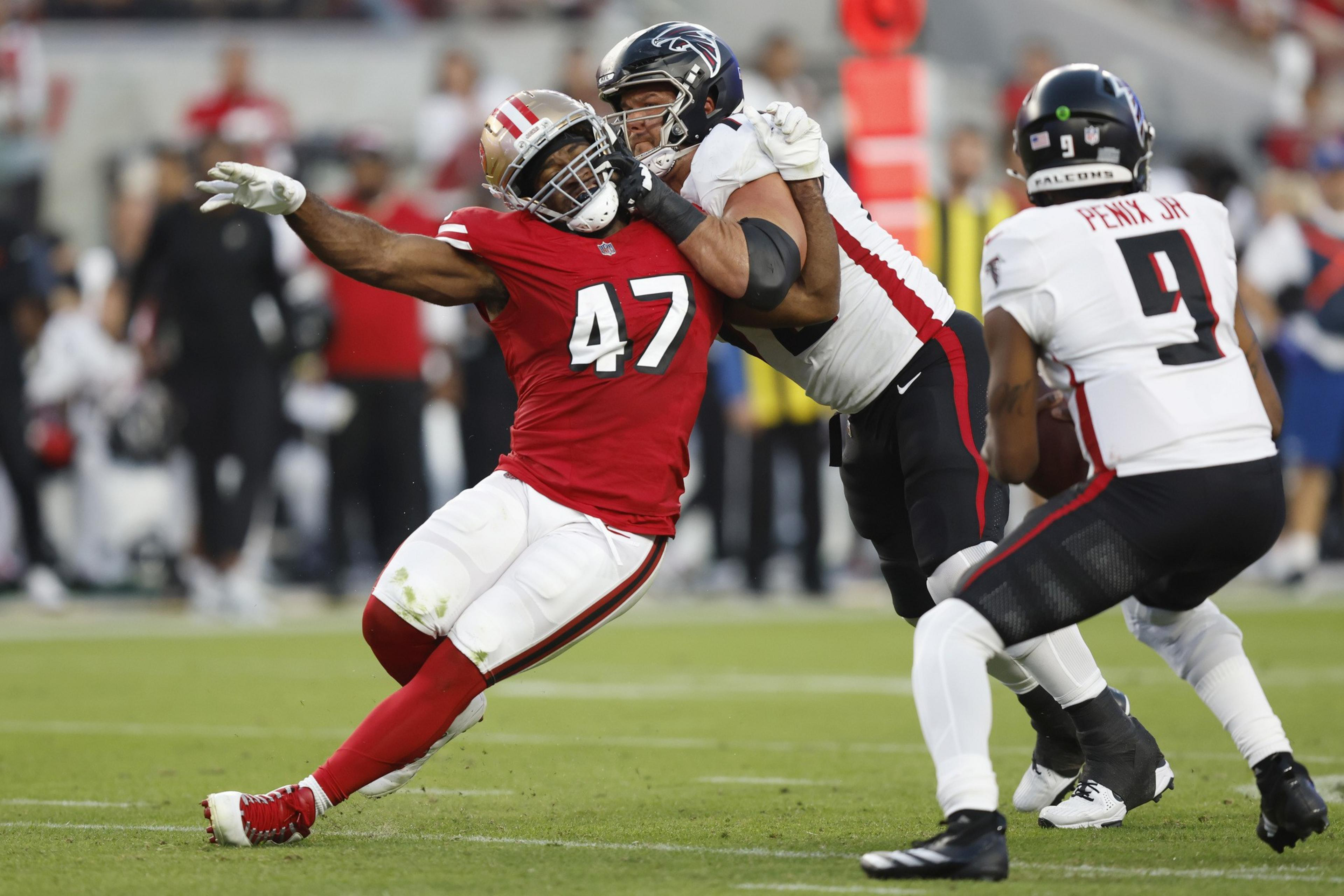 A San Francisco 49ers player wearing number 47 is being blocked by an Atlanta Falcons player while the Falcons quarterback, number 9, prepares to throw the ball.