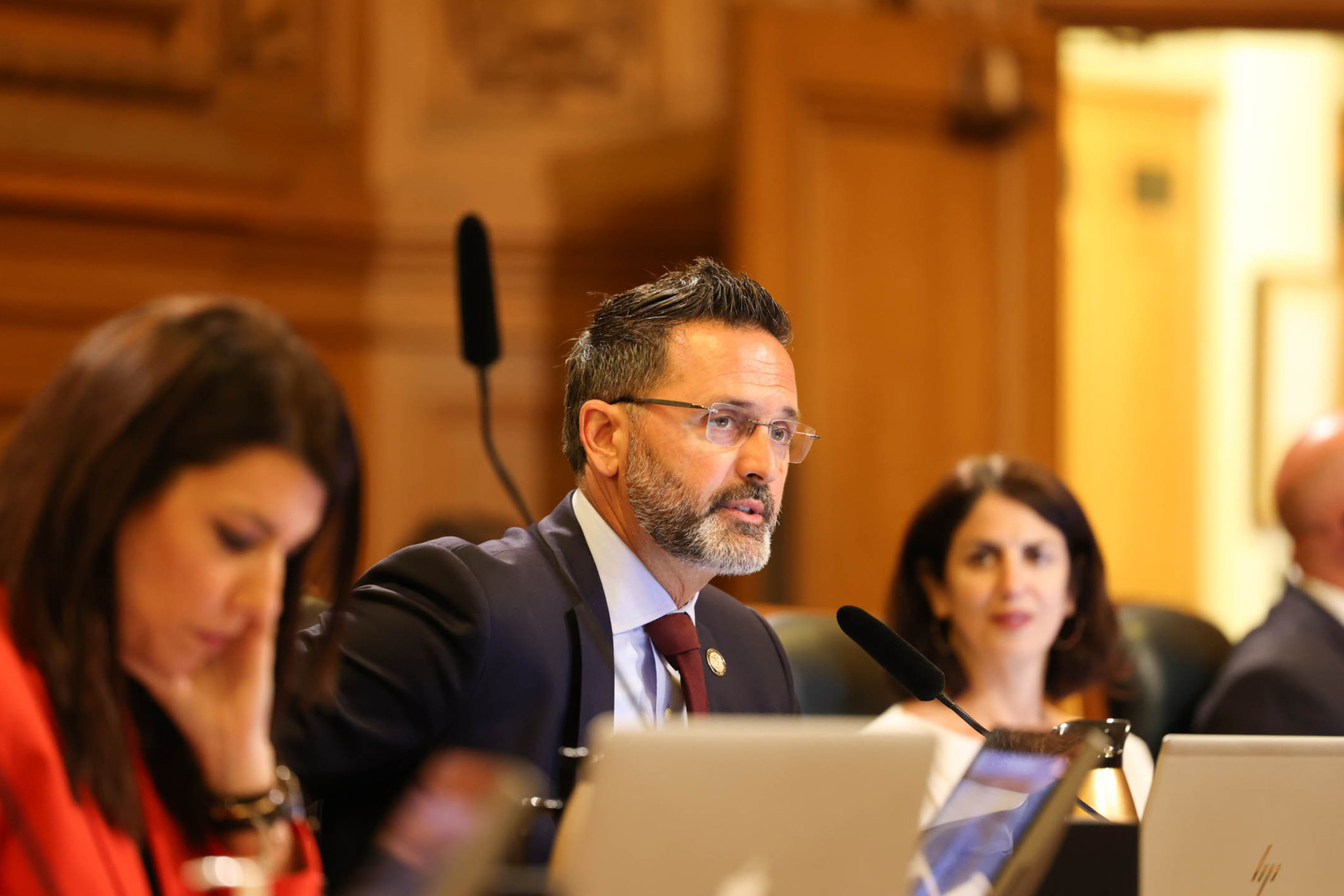 Supervisor Ahsha SafaÃ­, wearing glasses and a suit, participates in a formal meeting while two women colleagues sit on either side of him.