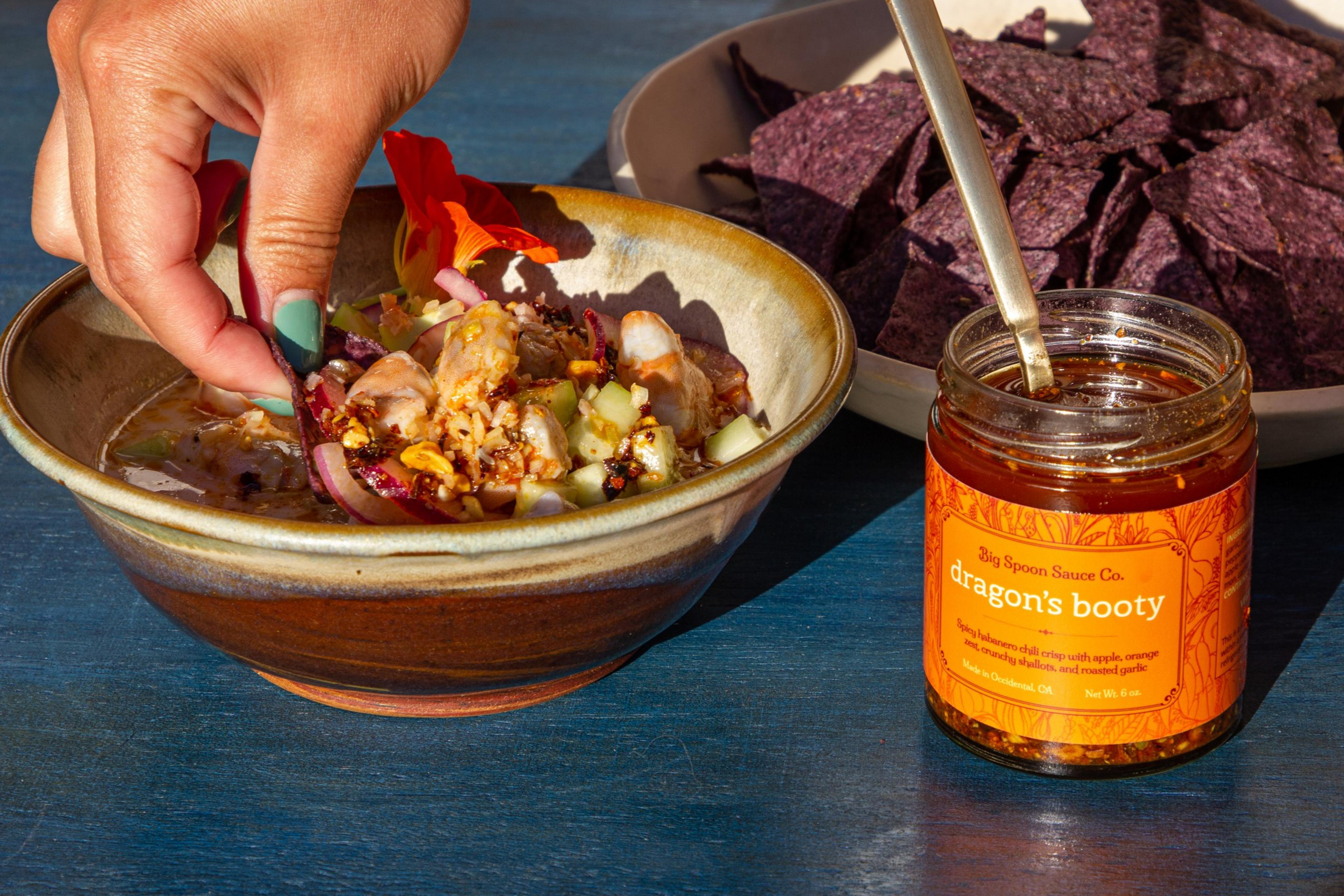 A jar of chili crisp sits on a table next to a bowl of salsa and chips. A hand dips a chip into the salsa.
