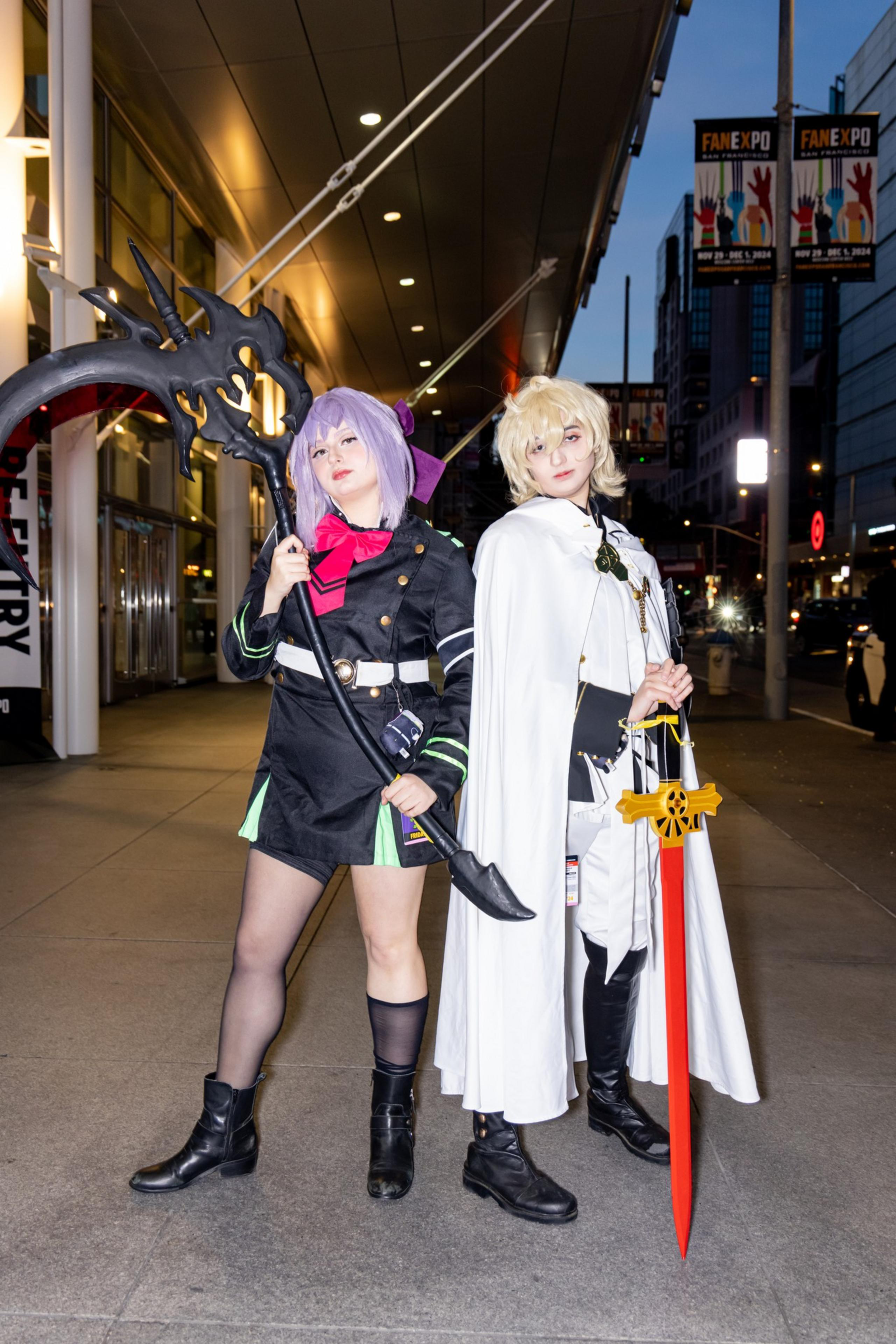 Two people in detailed cosplay costumes pose with weapons on a city street at night, under bright lights. One has purple hair, the other blonde.