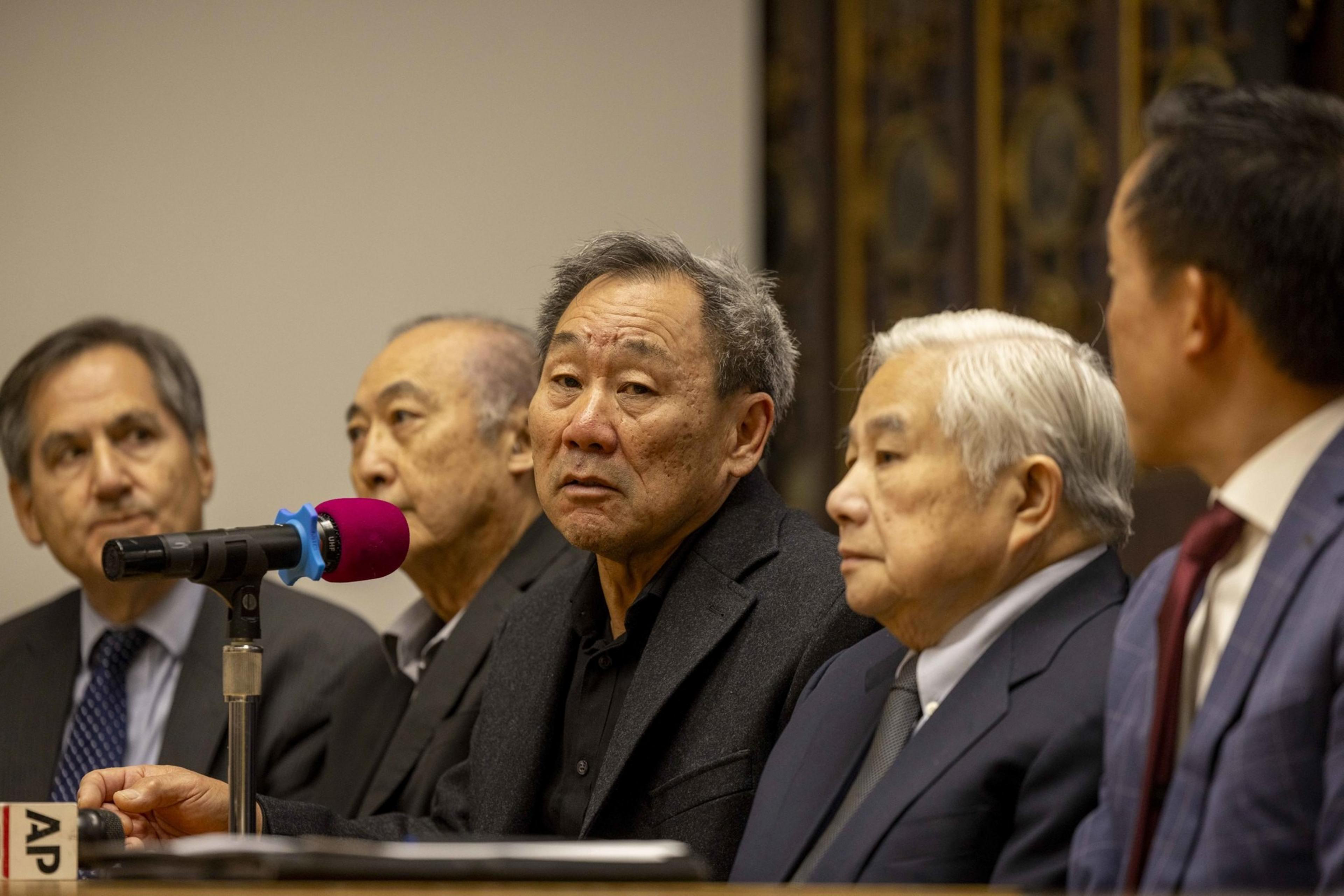Five men in suits sit at a panel with a microphone on the table. They appear serious and are focused on the speaker who is using the microphone.