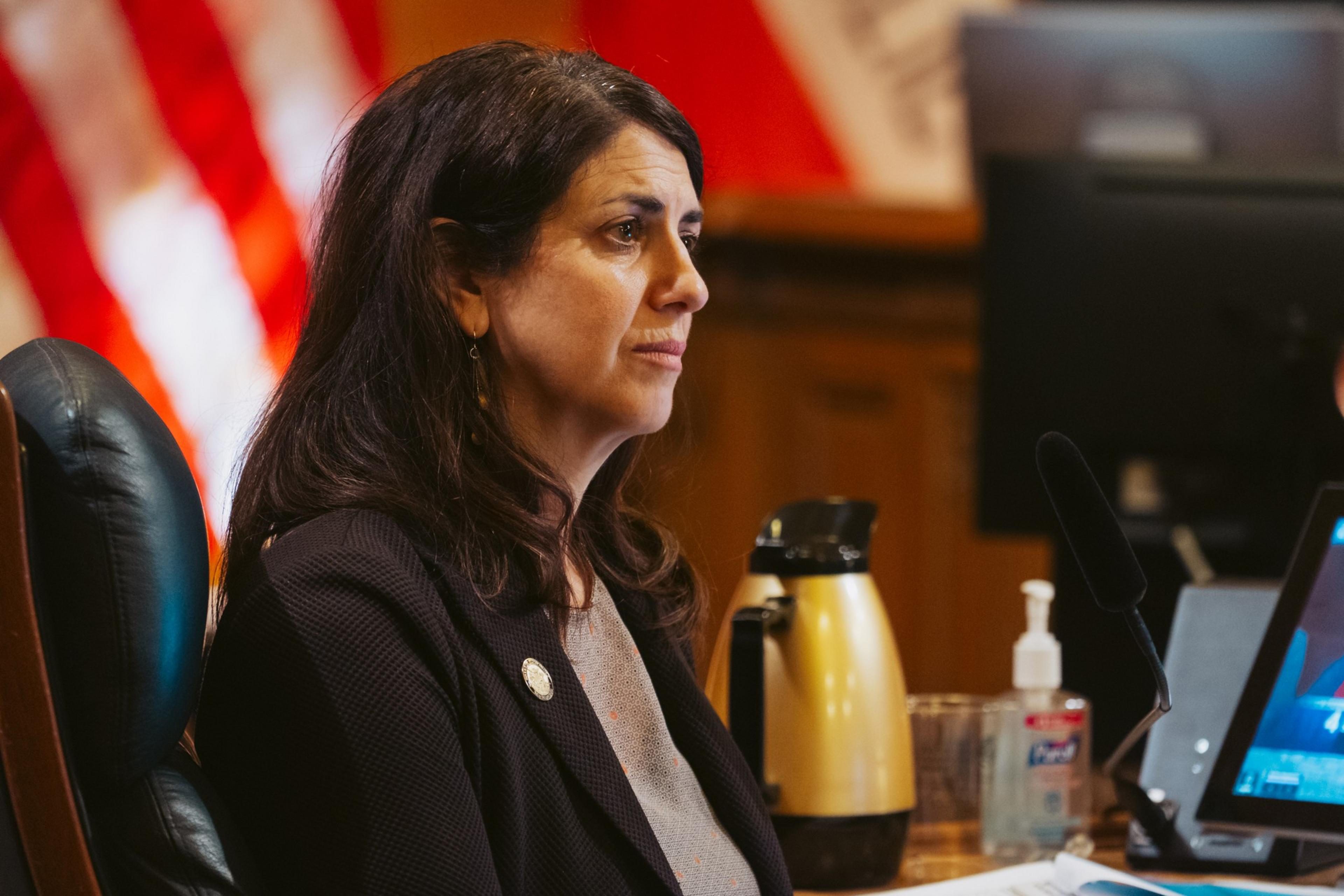 Hillary Ronen in a blazer listens as people talk in a room with an American flag in the background.