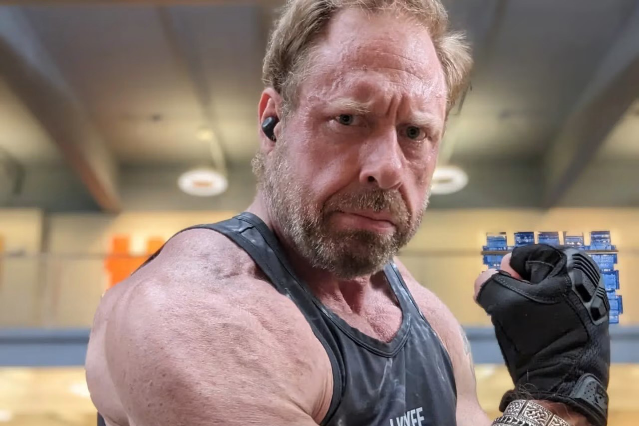 A muscular man wearing a black tank top flexes his arm, showing veins and wearing a fingerless glove and silver bracelets in a gym setting.