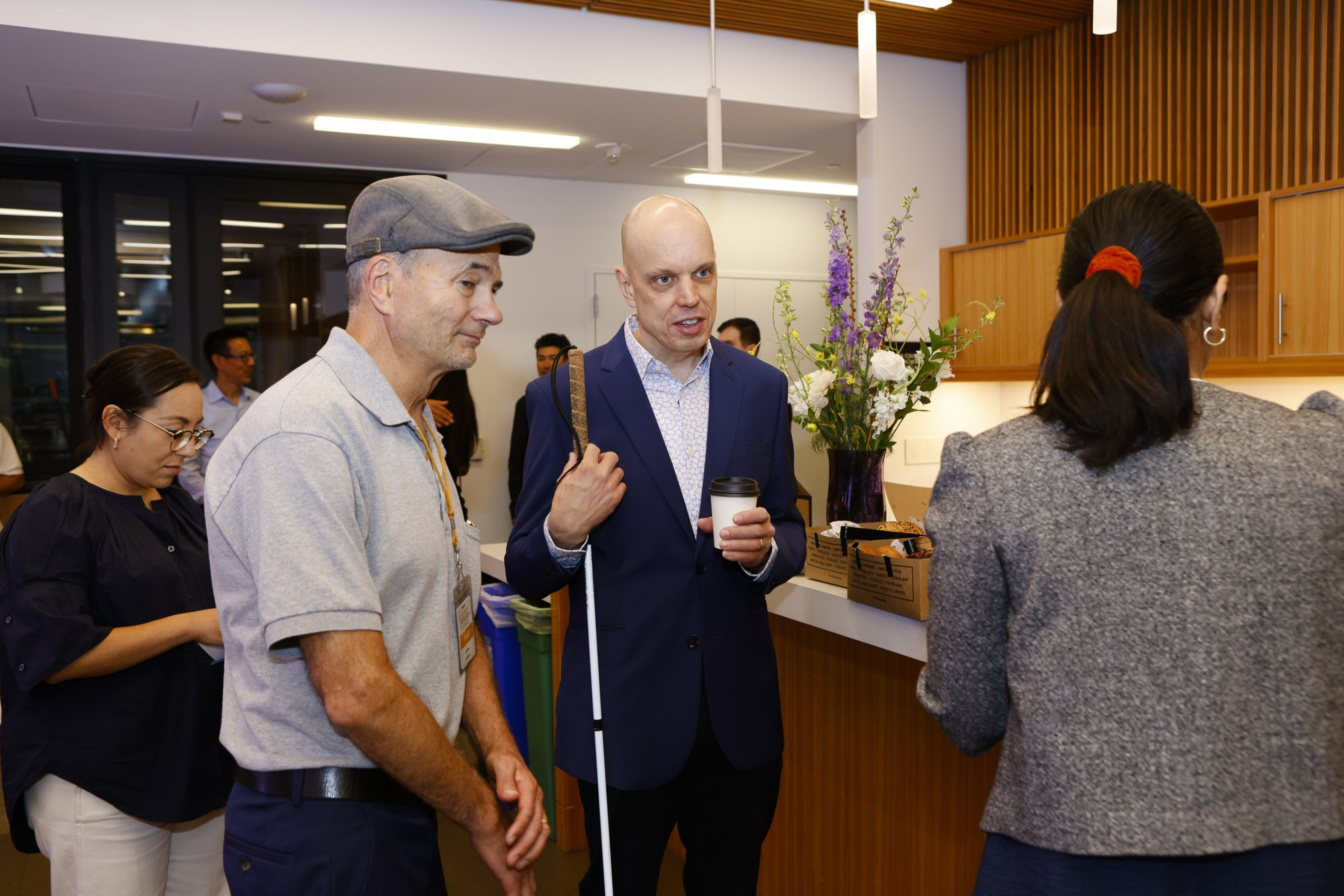 In a room with wooden cabinets and flowers, a man holding a white cane chats with a woman and another man wearing glasses, while people stand in the background.