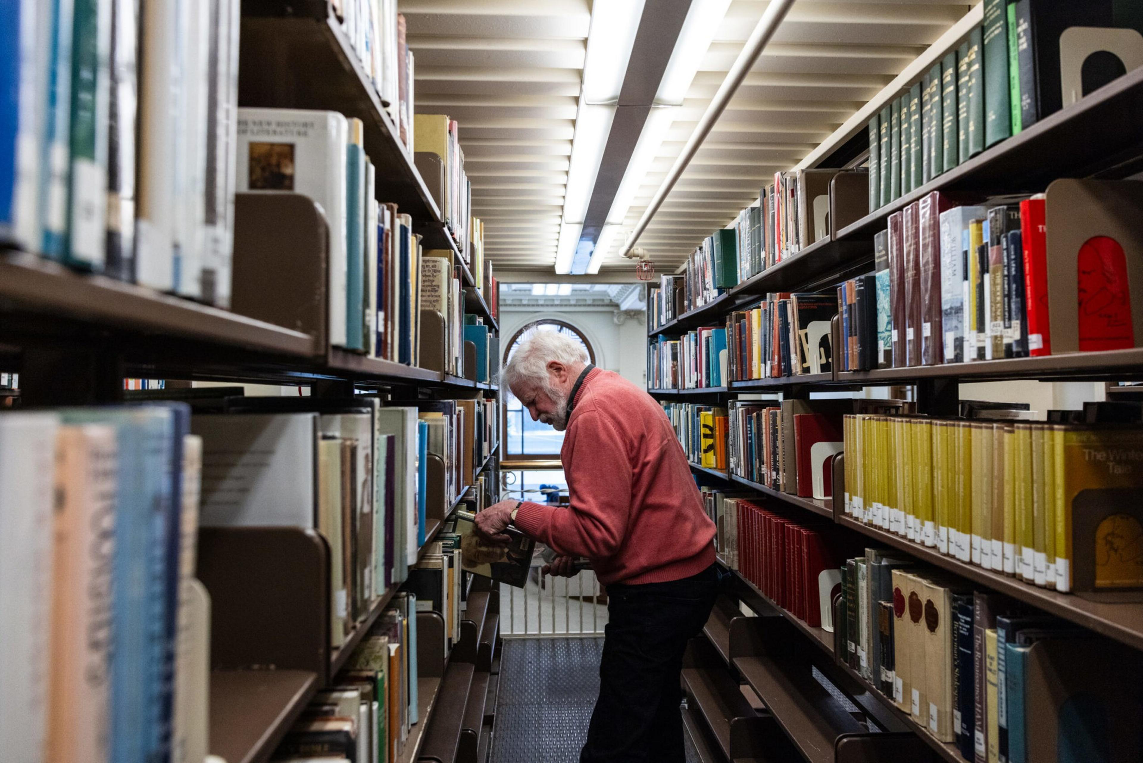 An elderly man in a red sweater searches through books on a shelf in a narrow, well-lit library aisle.