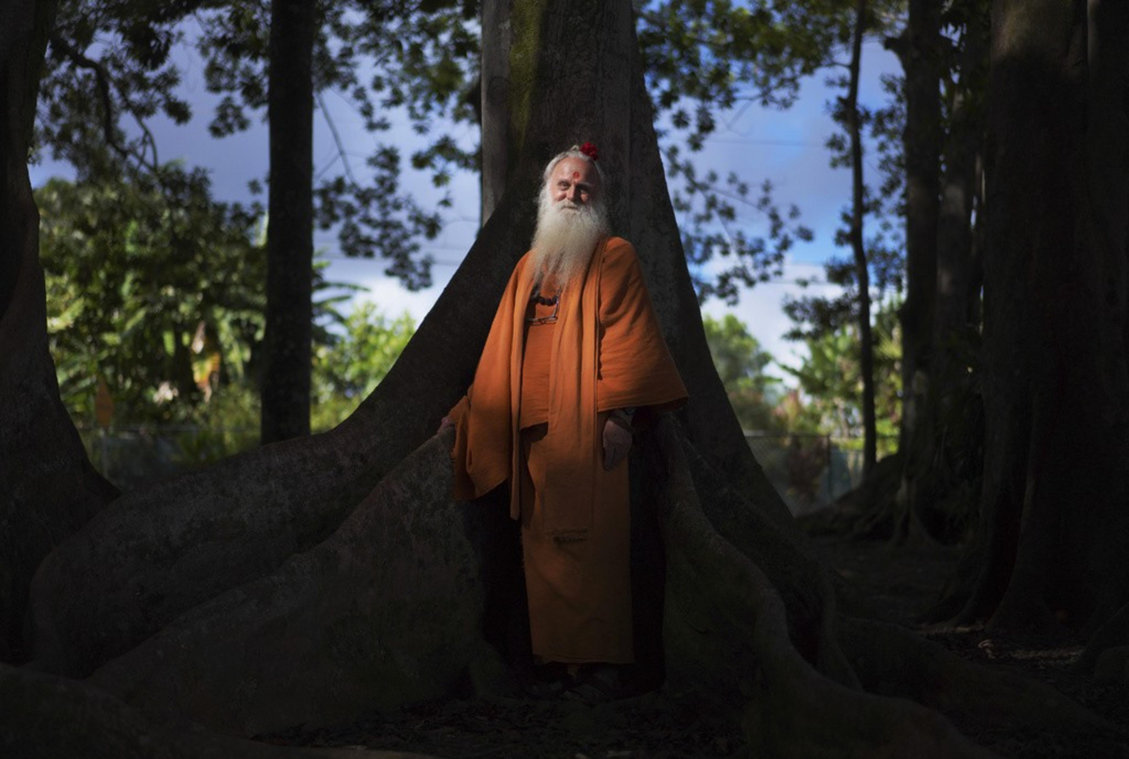 A man with a white beard in an orange robe sits in front of tall trees. 