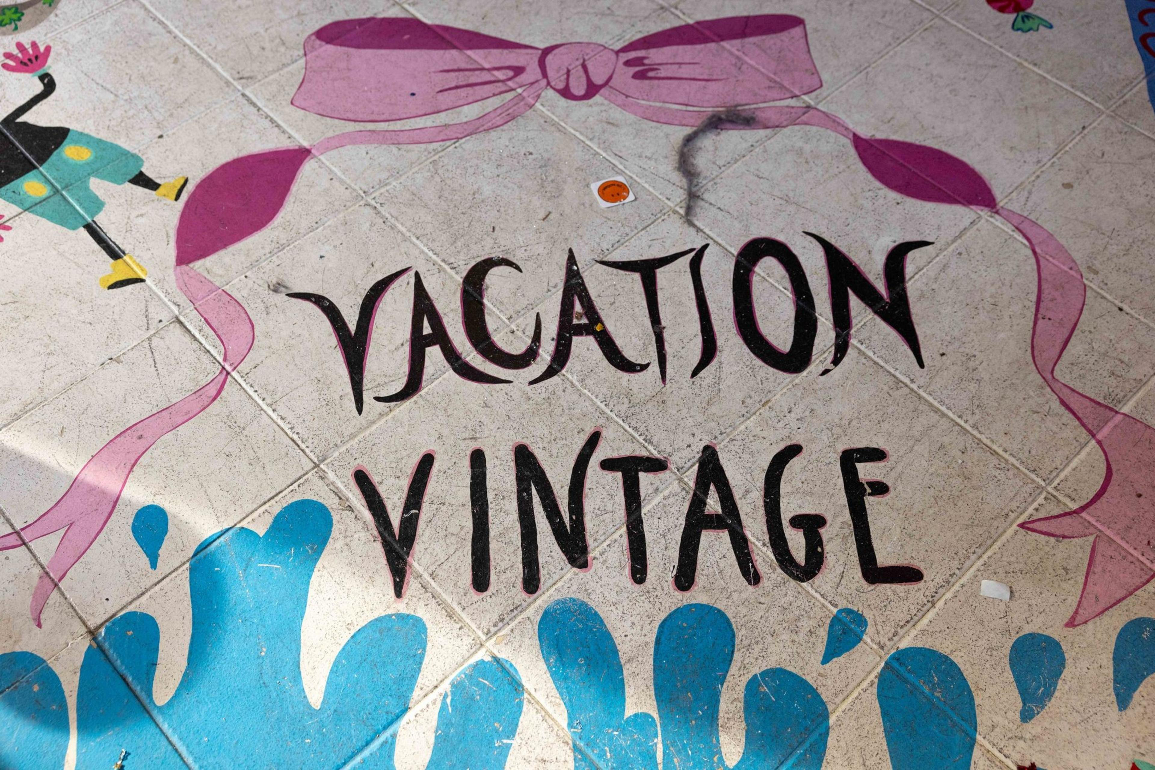 The image shows a tiled floor with &quot;Vacation Vintage&quot; in bold letters, framed by a pink bow at the top and blue waves at the bottom.