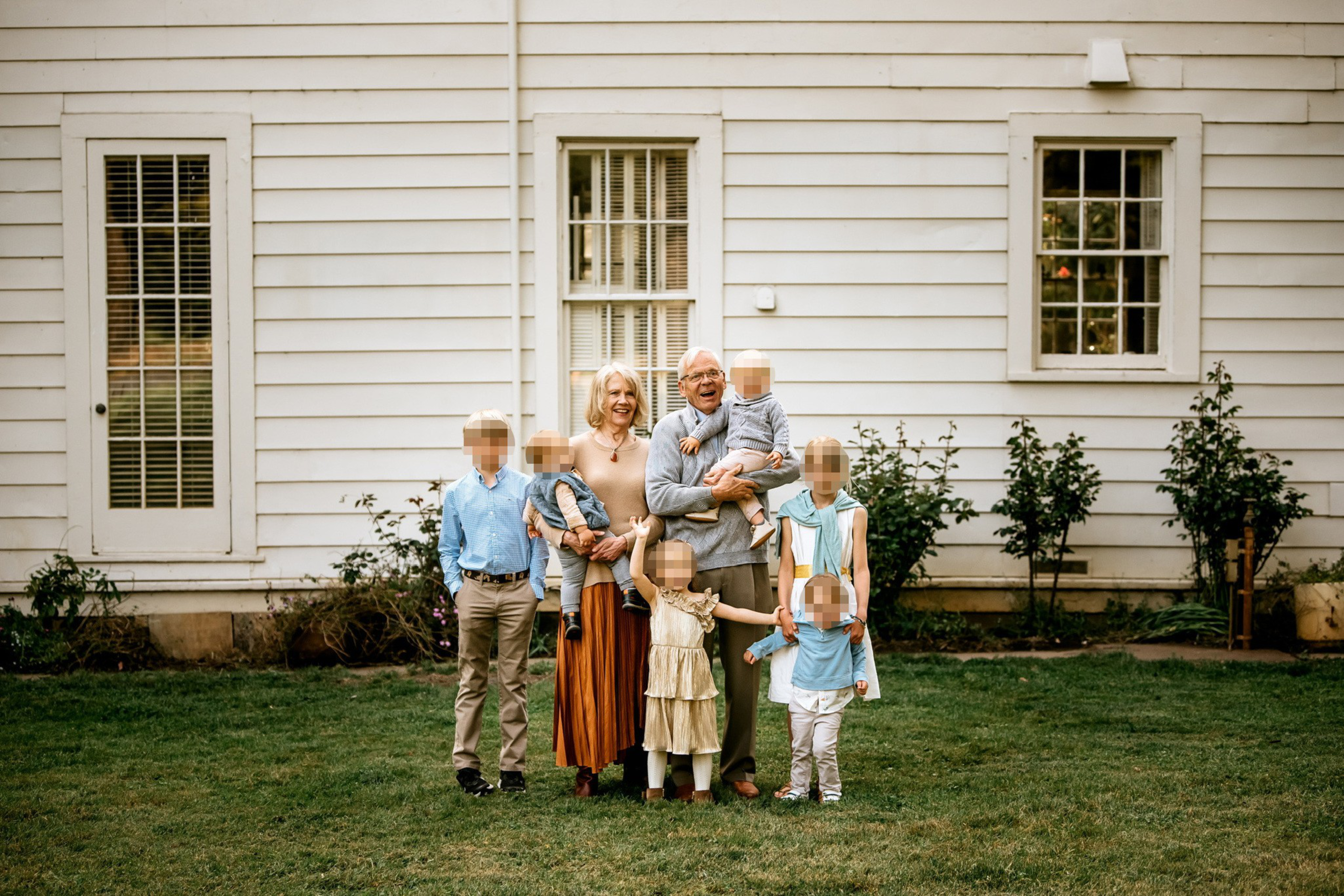 A group of seven people stands in front of a white house. An older couple is in the center, surrounded by children, with greenery and two windows in the background.