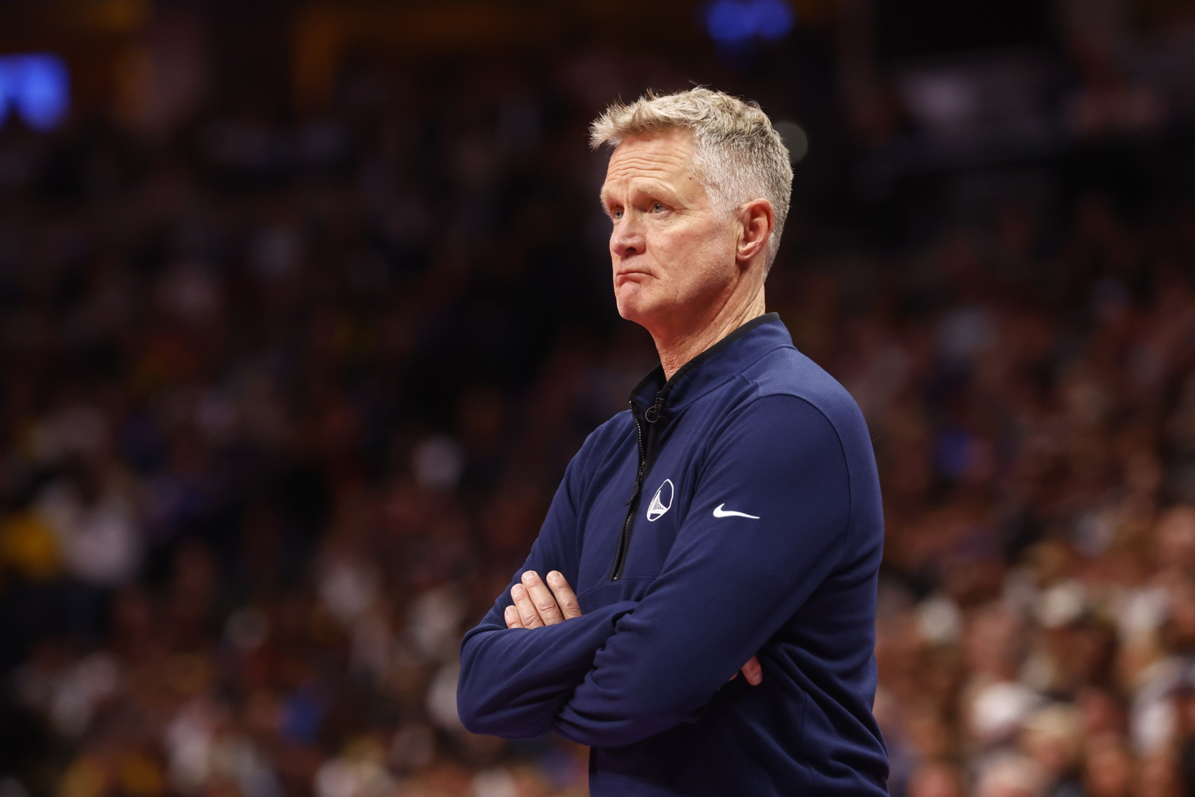A man with short blond hair wearing a navy blue Golden State Warriors jacket stands with his arms crossed, looking thoughtfully to the side.