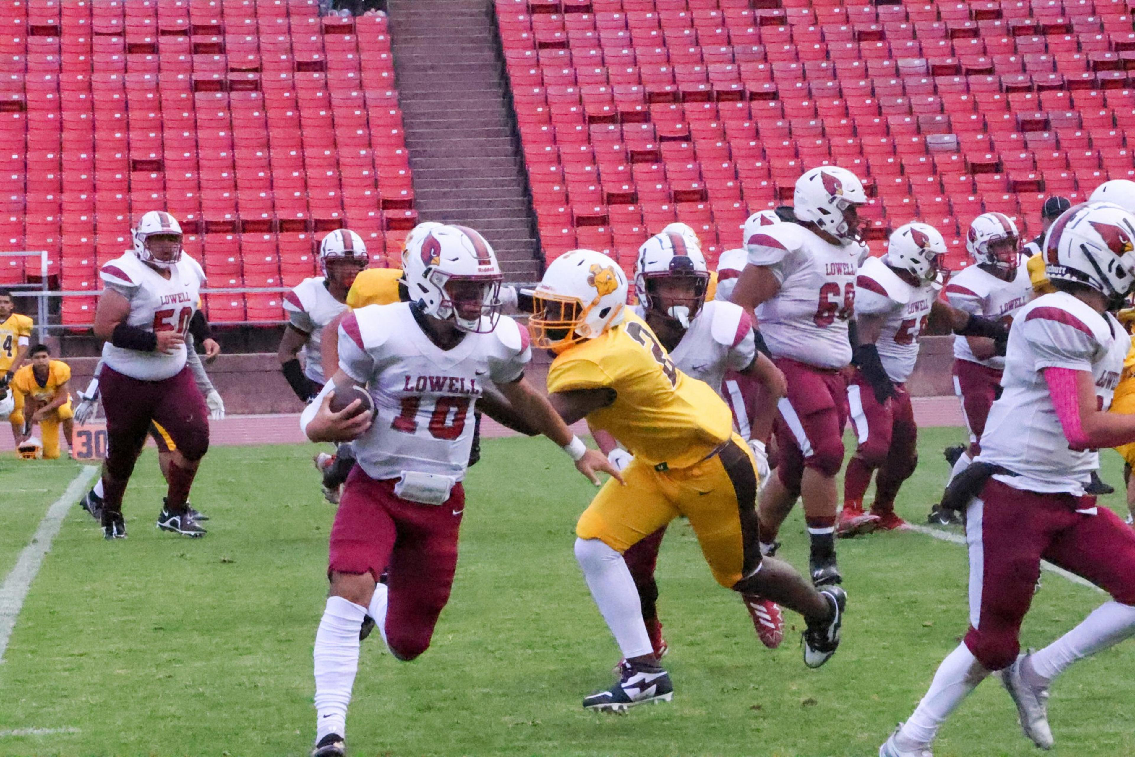 A football player in a white and maroon Lowell uniform carries the ball while being chased by a player in a yellow uniform on a green field with empty red stadium seats.