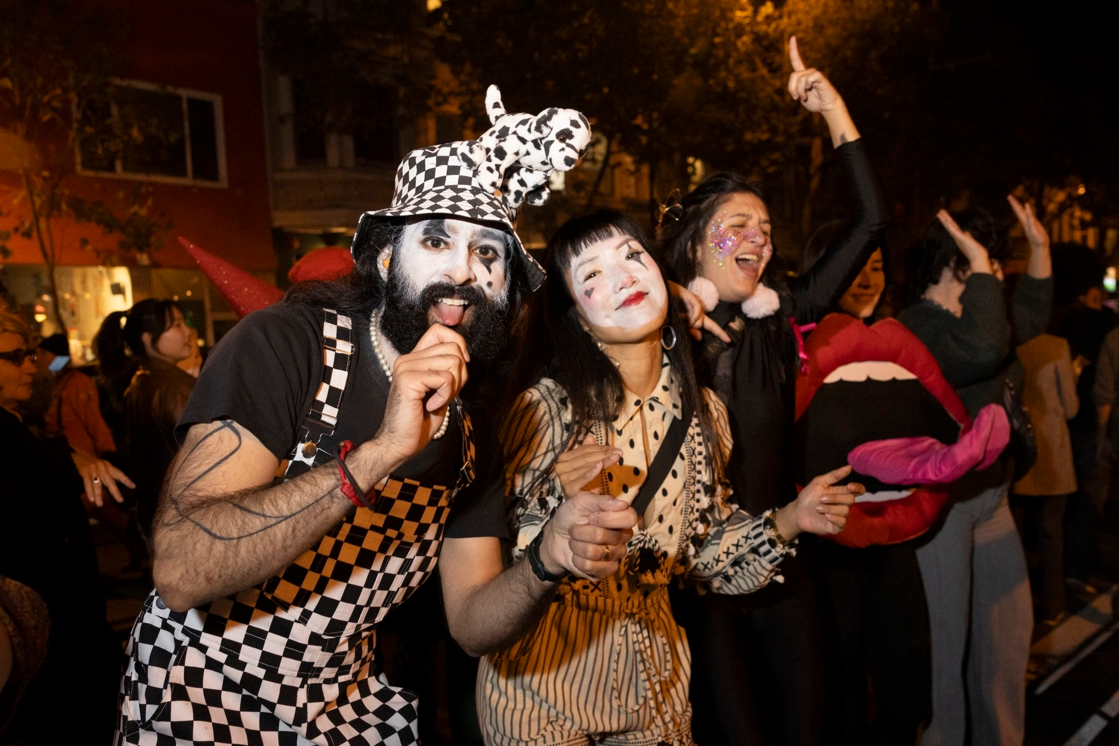 Three people in costumes and face paint are celebrating at night. One is in checkered attire, another in a striped blouse, and the third with glitter and a playful prop.