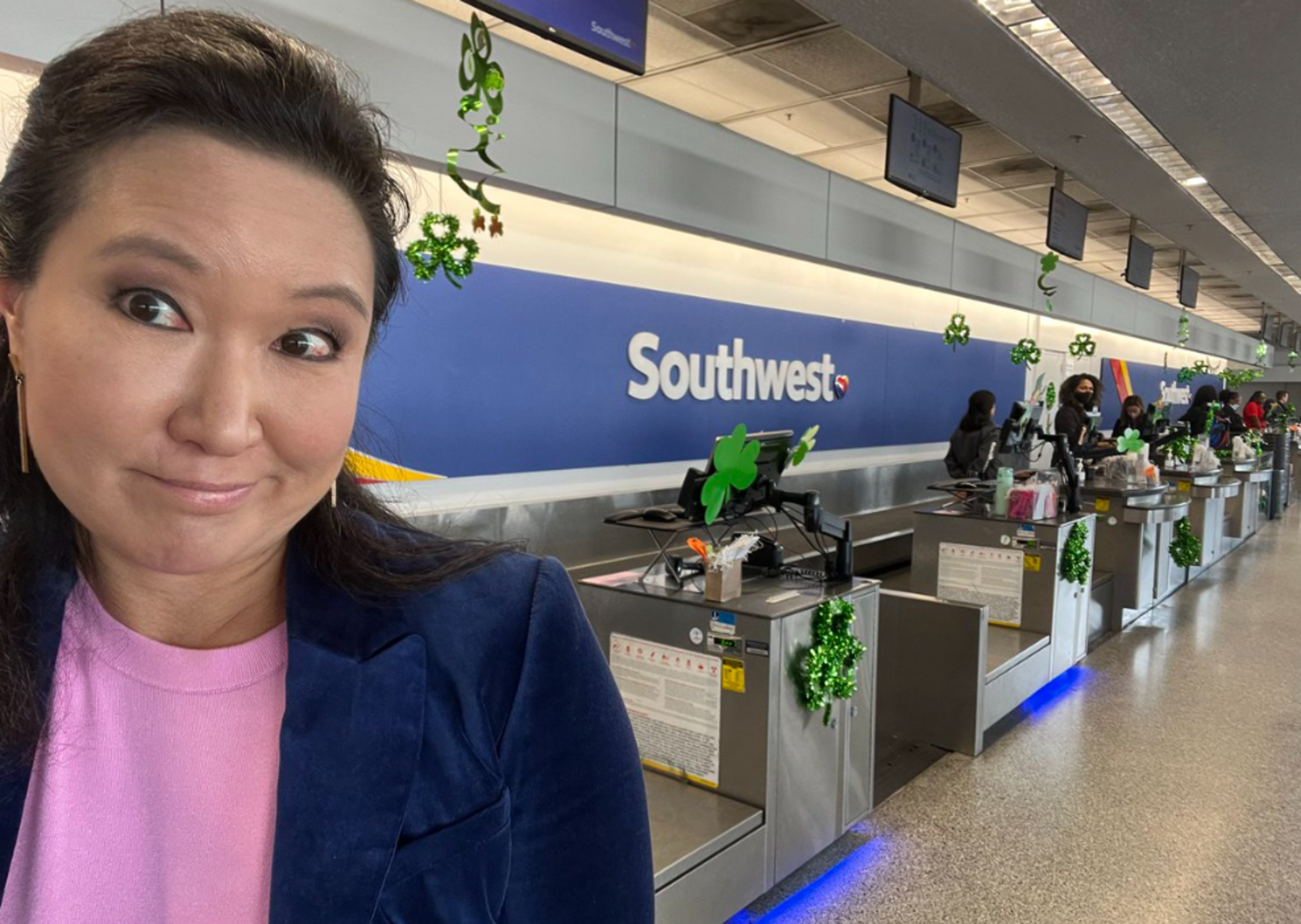A woman in a pink shirt and blue jacket stands near a Southwest Airlines check-in counter decorated with green shamrocks for St. Patrick's Day.