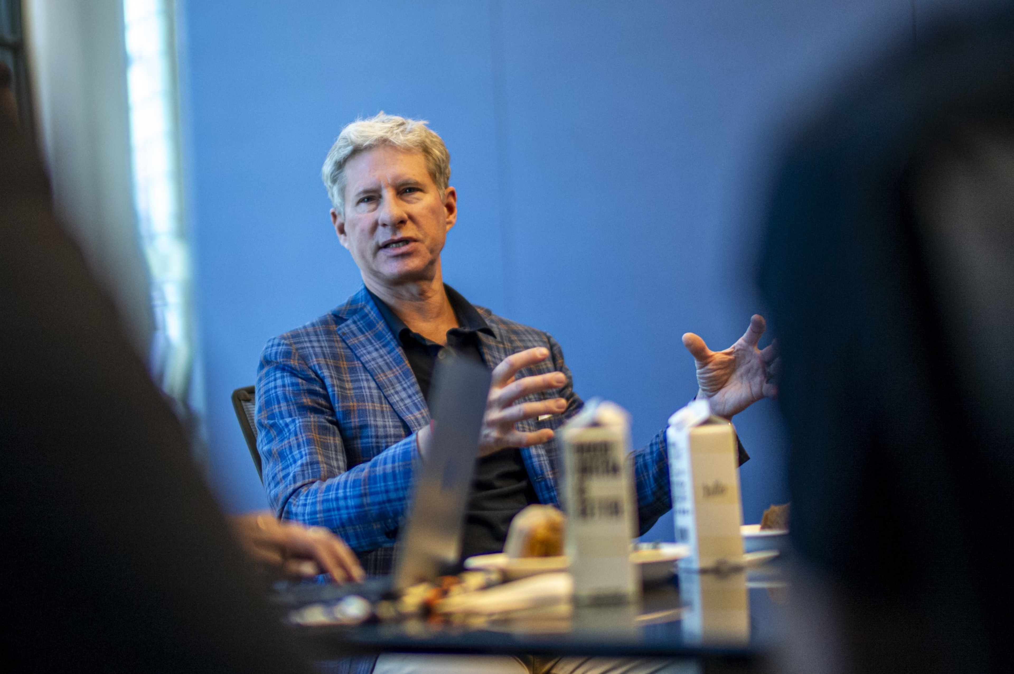 A man in a checkered blazer gestures while speaking in a meeting, with bottled drinks and a blurred foreground.