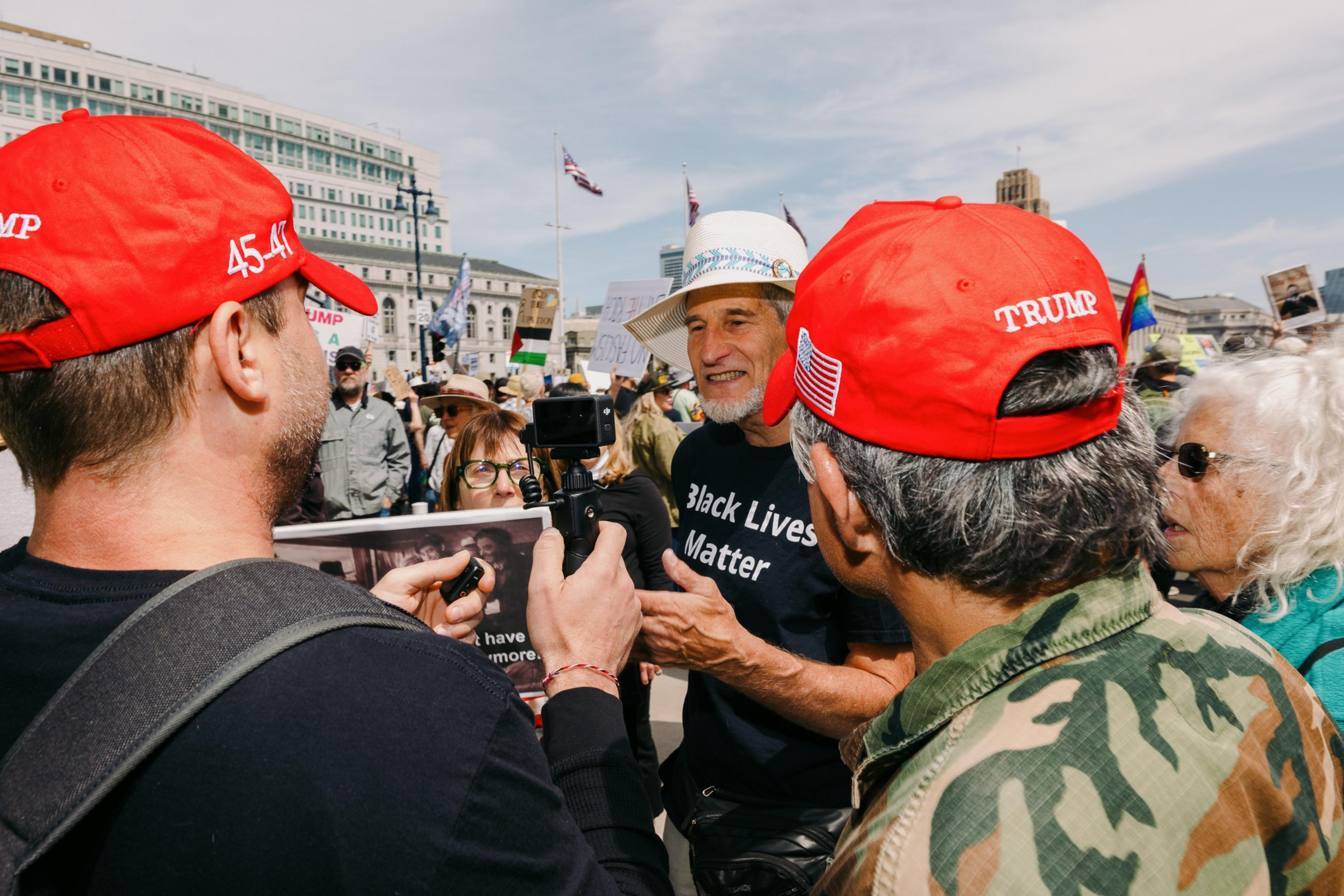A group of people are having a conversation at a protest. One wears a &quot;Black Lives Matter&quot; shirt, while others wear red caps with &quot;Trump&quot; on them. Signs and flags are visible.