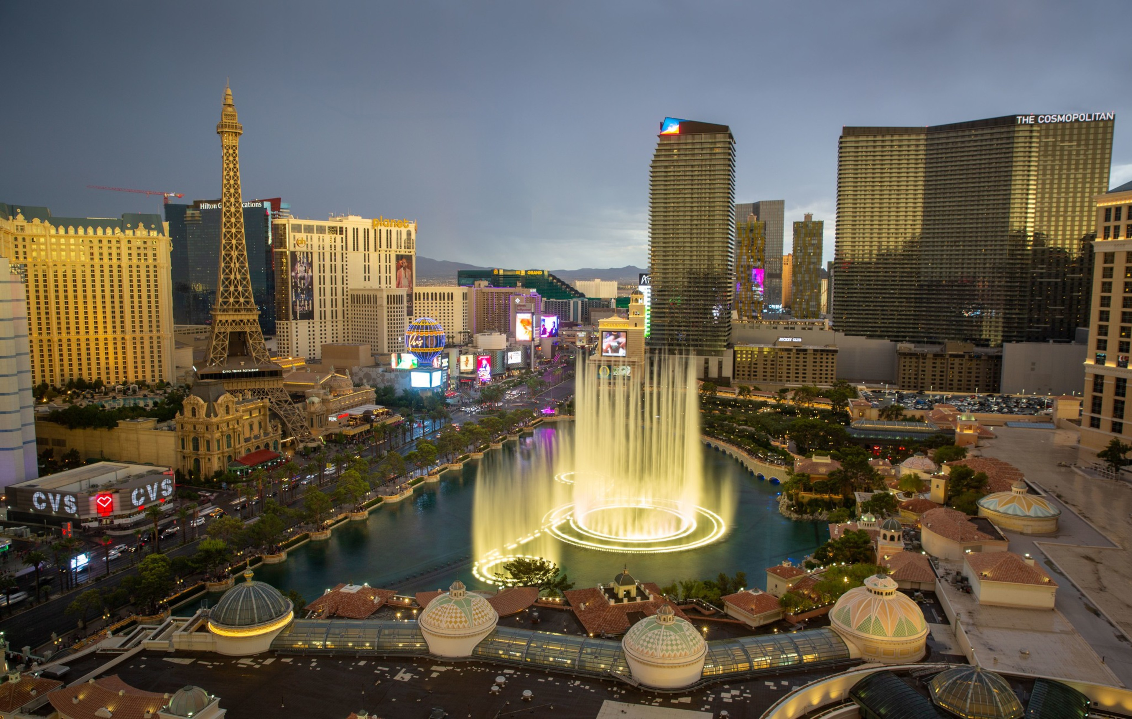 The image shows a bustling cityscape with tall buildings, a smaller replica of the Eiffel Tower, and a large fountain illuminated by lights at dusk.