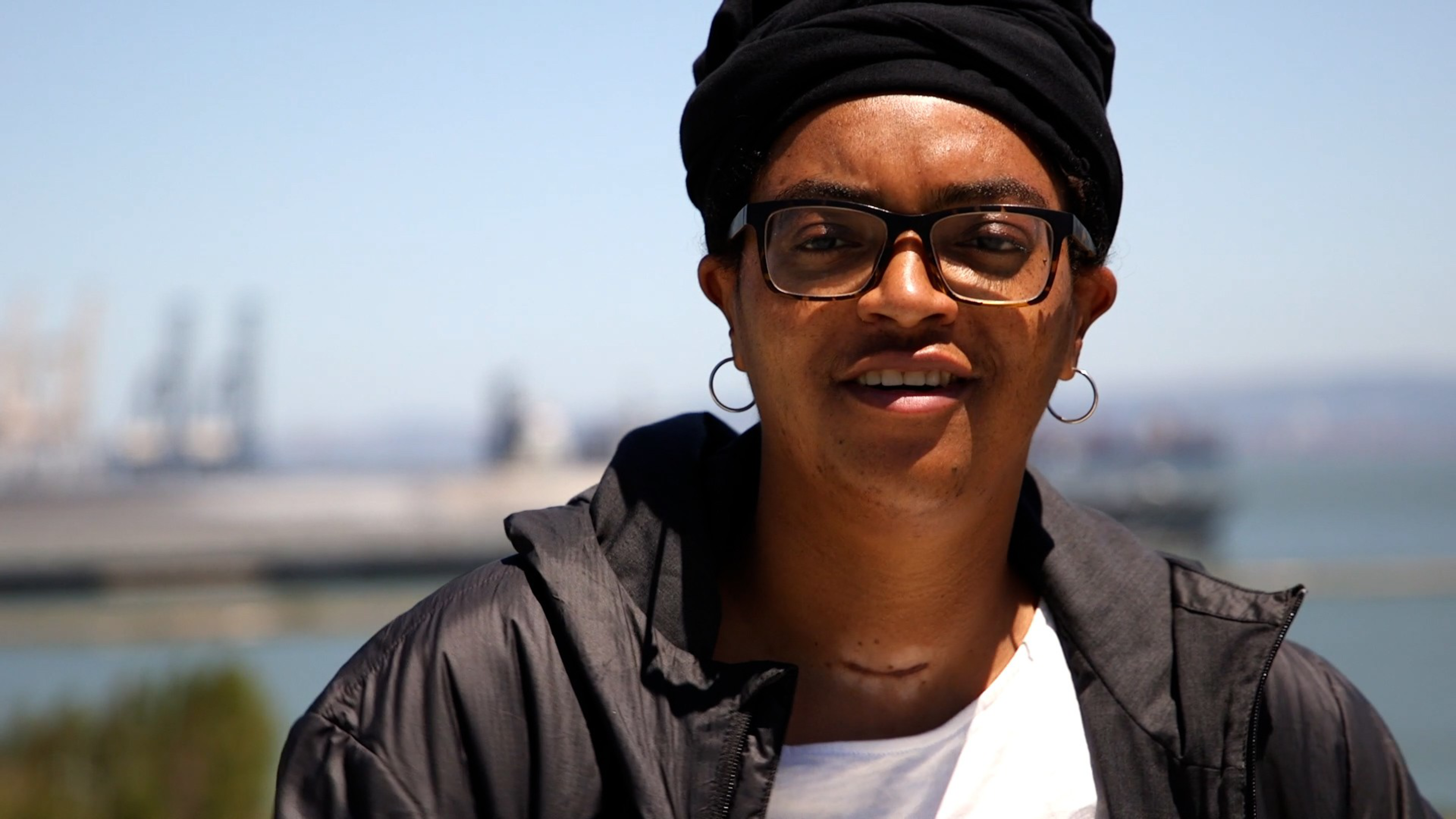 A person wearing glasses, a black head wrap, hoop earrings, a black jacket, and a white shirt stands outdoors near a body of water with blurred industrial structures in the background.
