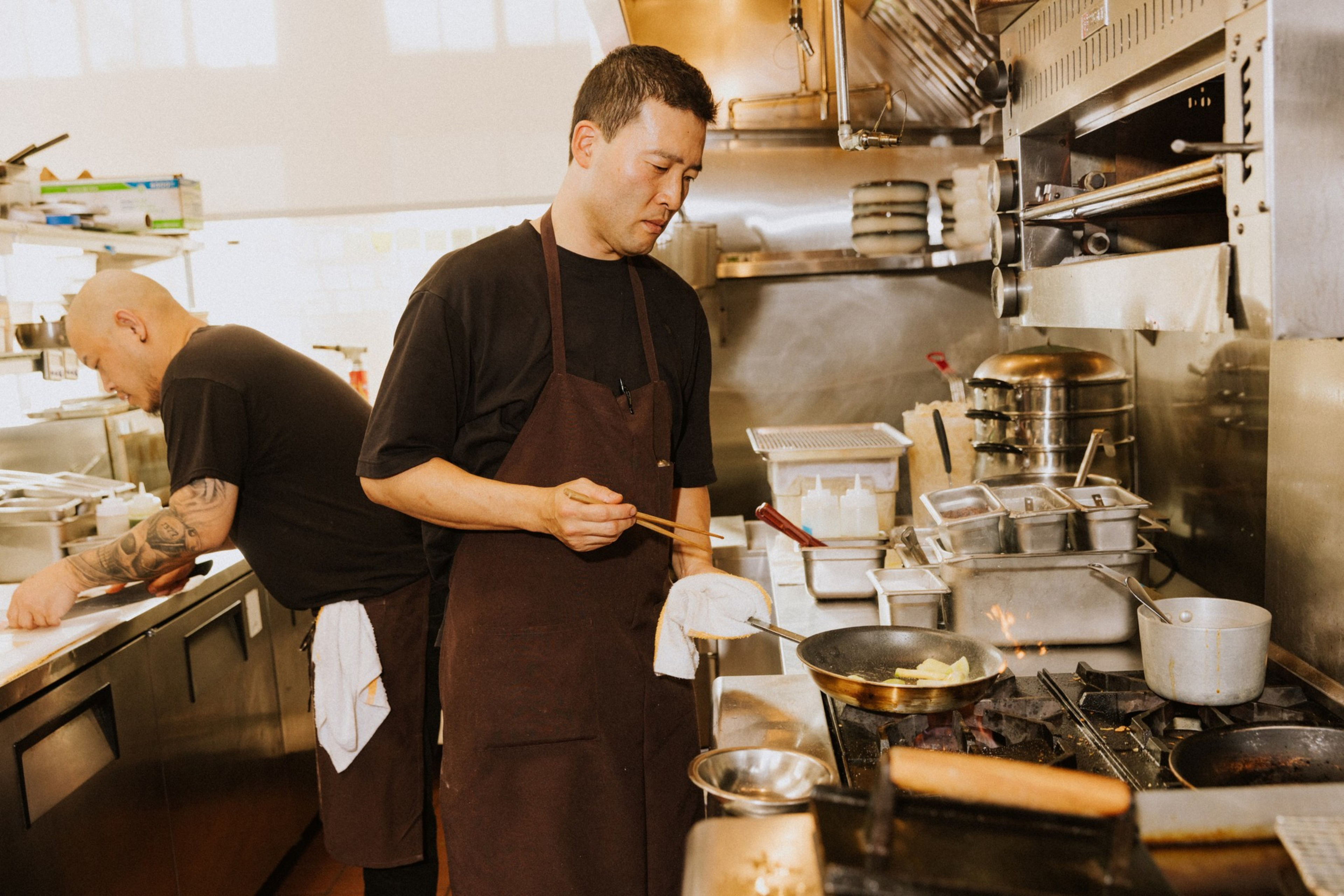 Two chefs in a kitchen; one is cooking with a frying pan on a stove, using chopsticks, while the other is preparing food at a counter. Stainless steel equipment surrounds them.