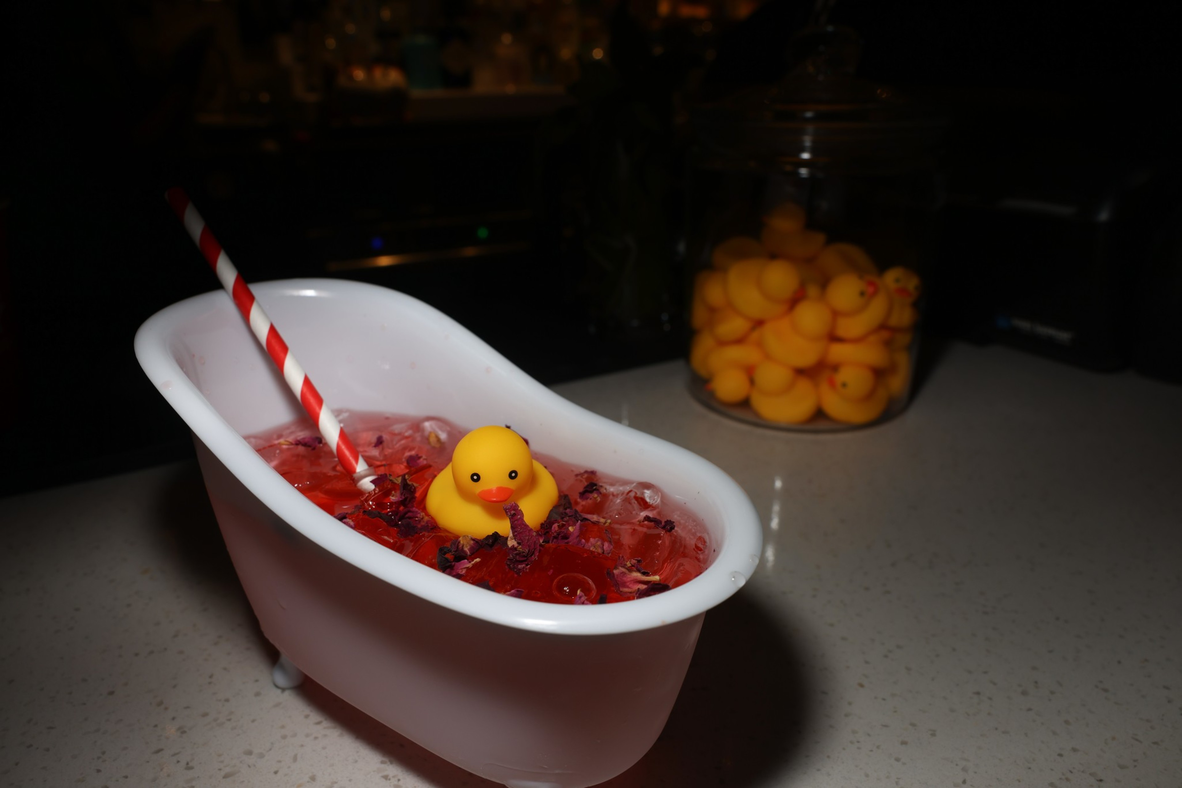 A small white bathtub-shaped container holds a red drink with ice, a striped straw, and a rubber duck; in the background, there is a jar filled with rubber ducks.