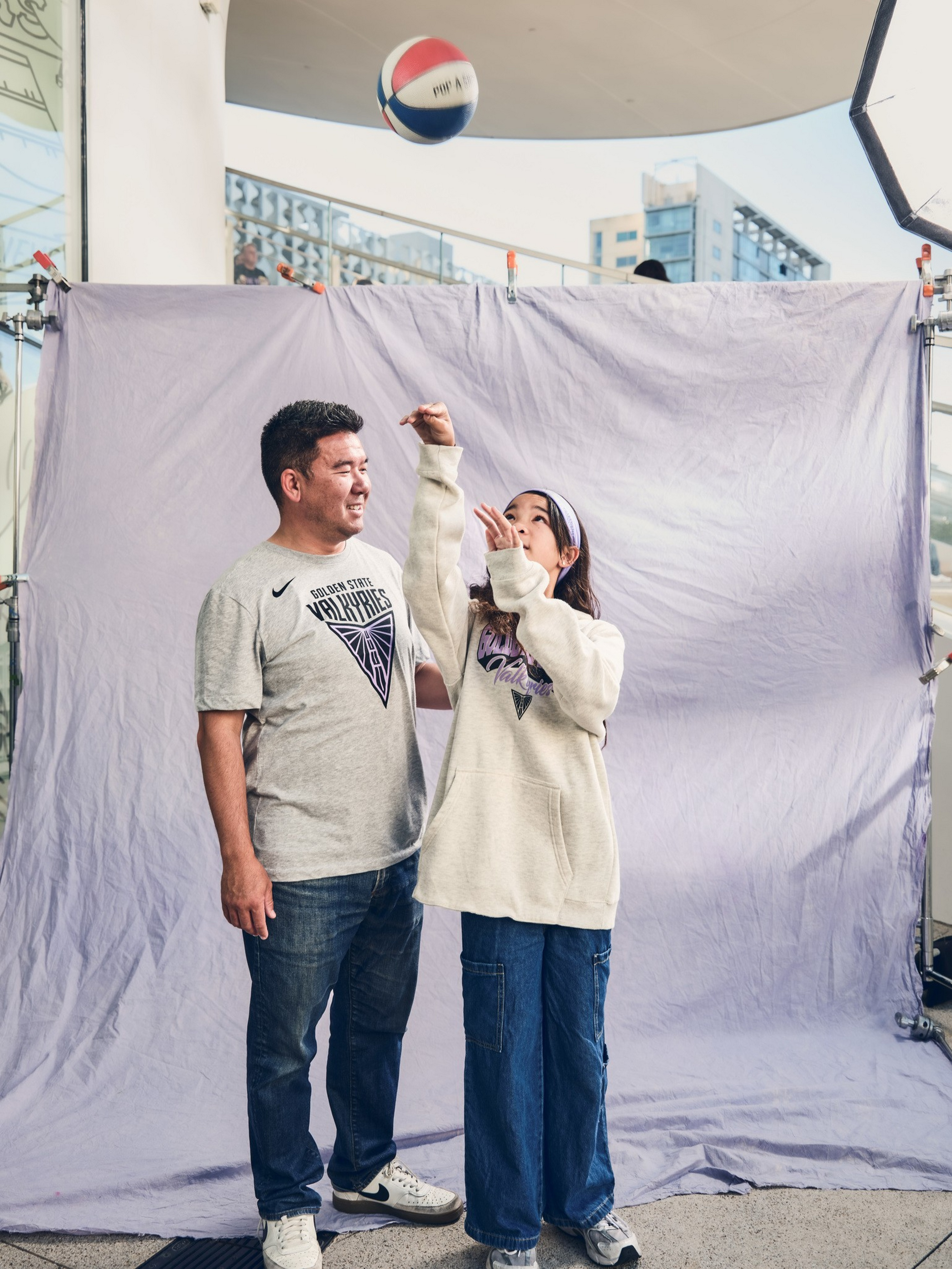 A man and a young girl stand in front of a fabric backdrop, wearing Golden State Valkyries shirts, tossing a red, white, and blue basketball.