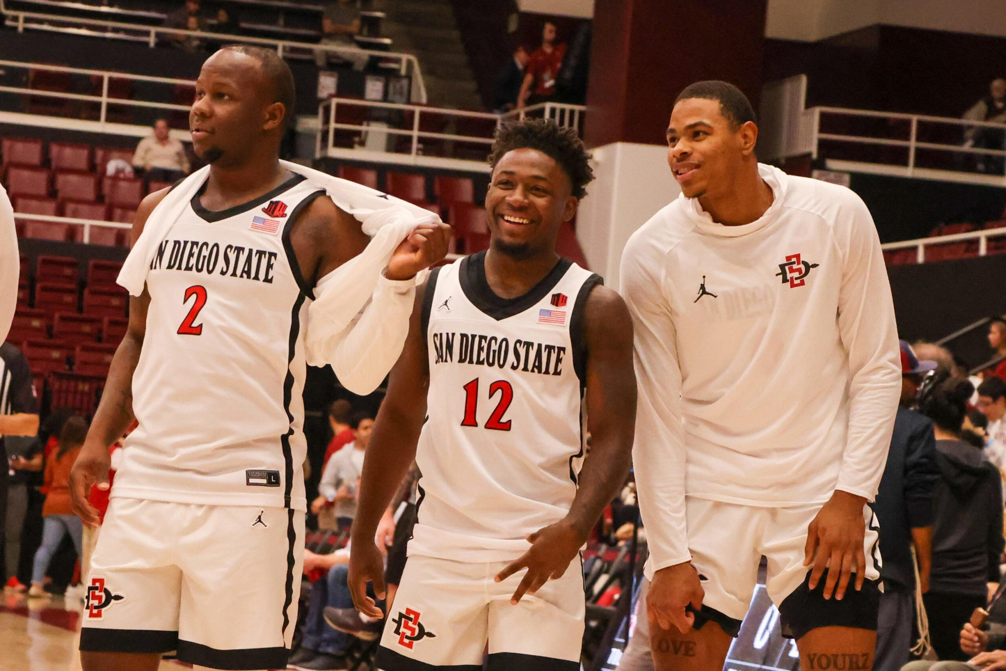 Three San Diego State basketball players, two in game uniforms and one in warm-up gear, stand smiling and relaxed on the court.