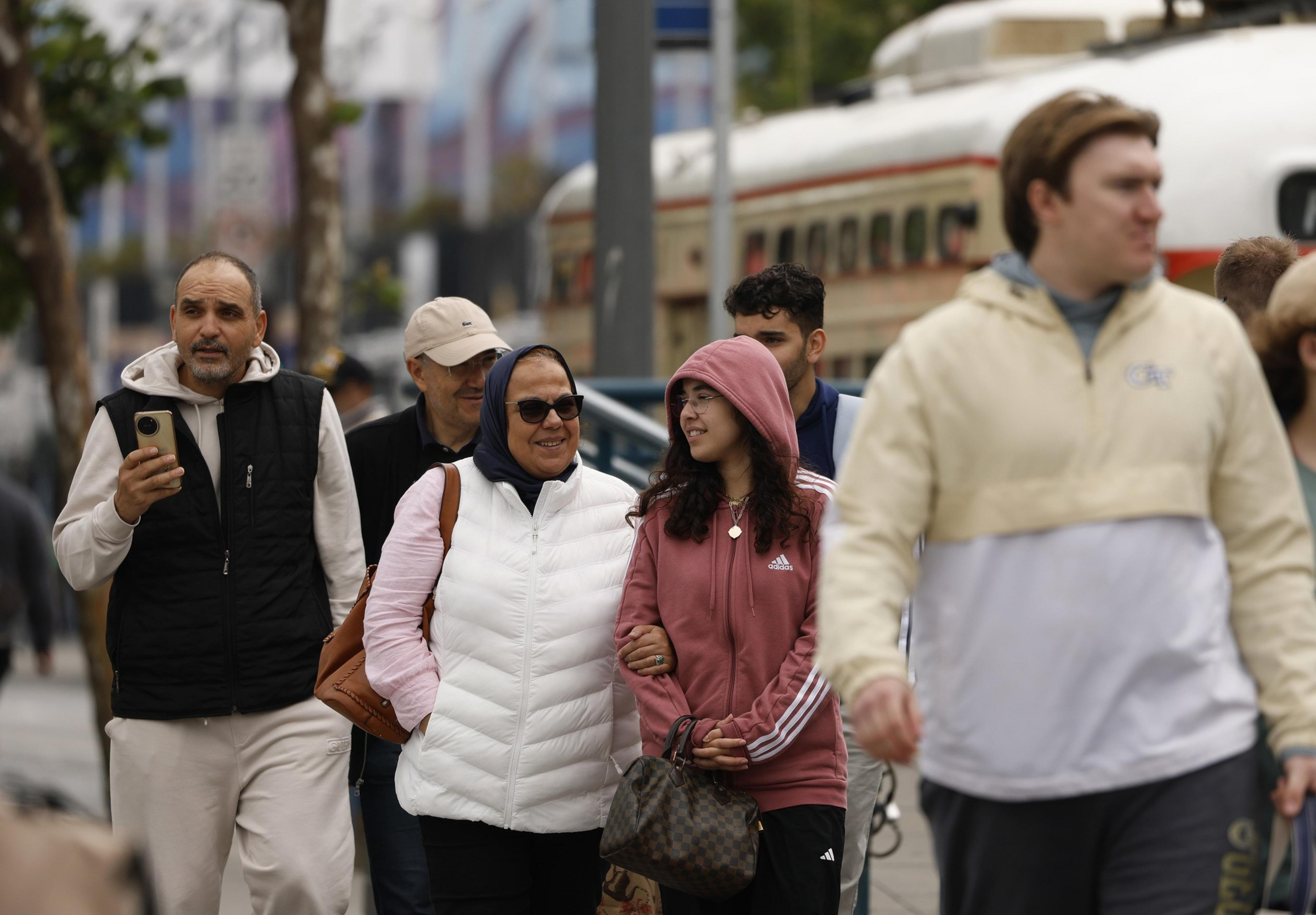 A group of people walks casually down a street. They wear casual clothing, and one person is holding a phone. A historic streetcar is in the background.