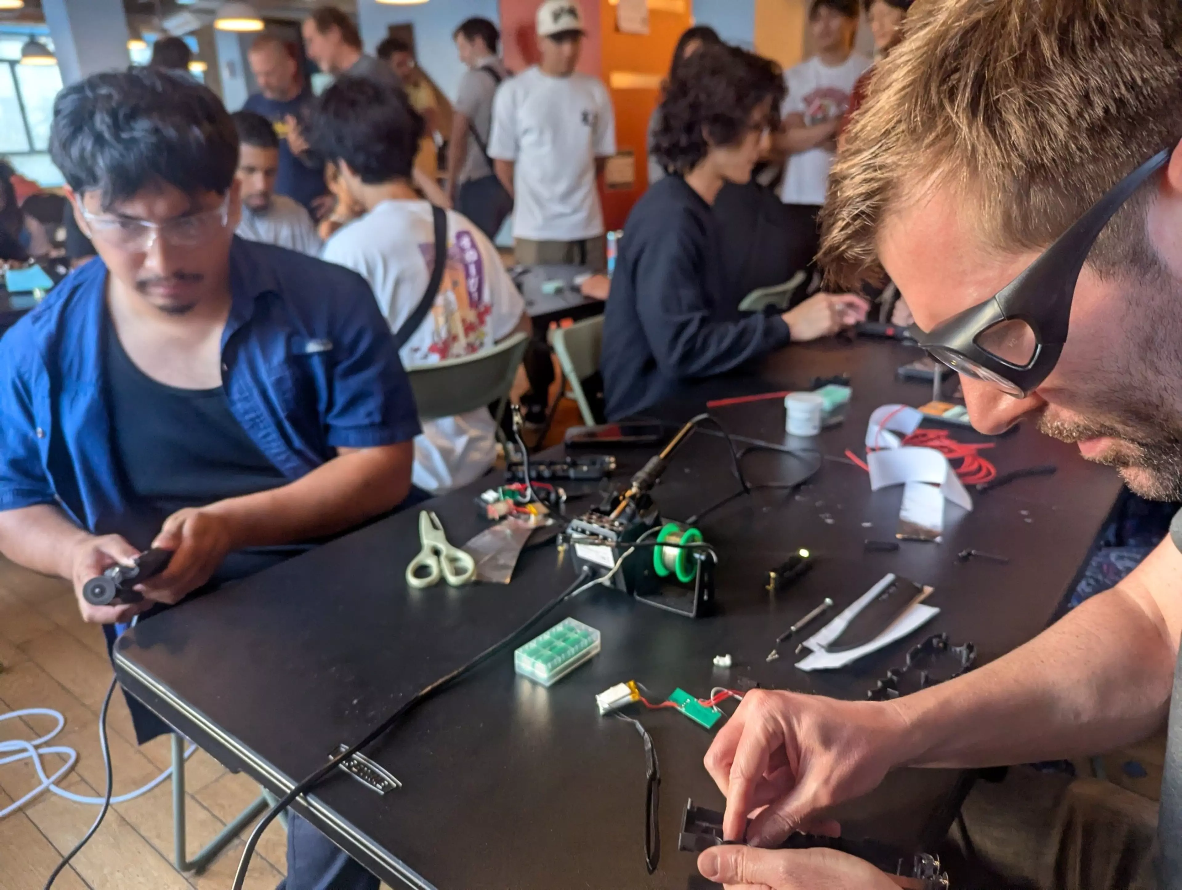 Two men work on electronic and mechanical components at a table cluttered with tools and wires, while several people talk in the background.