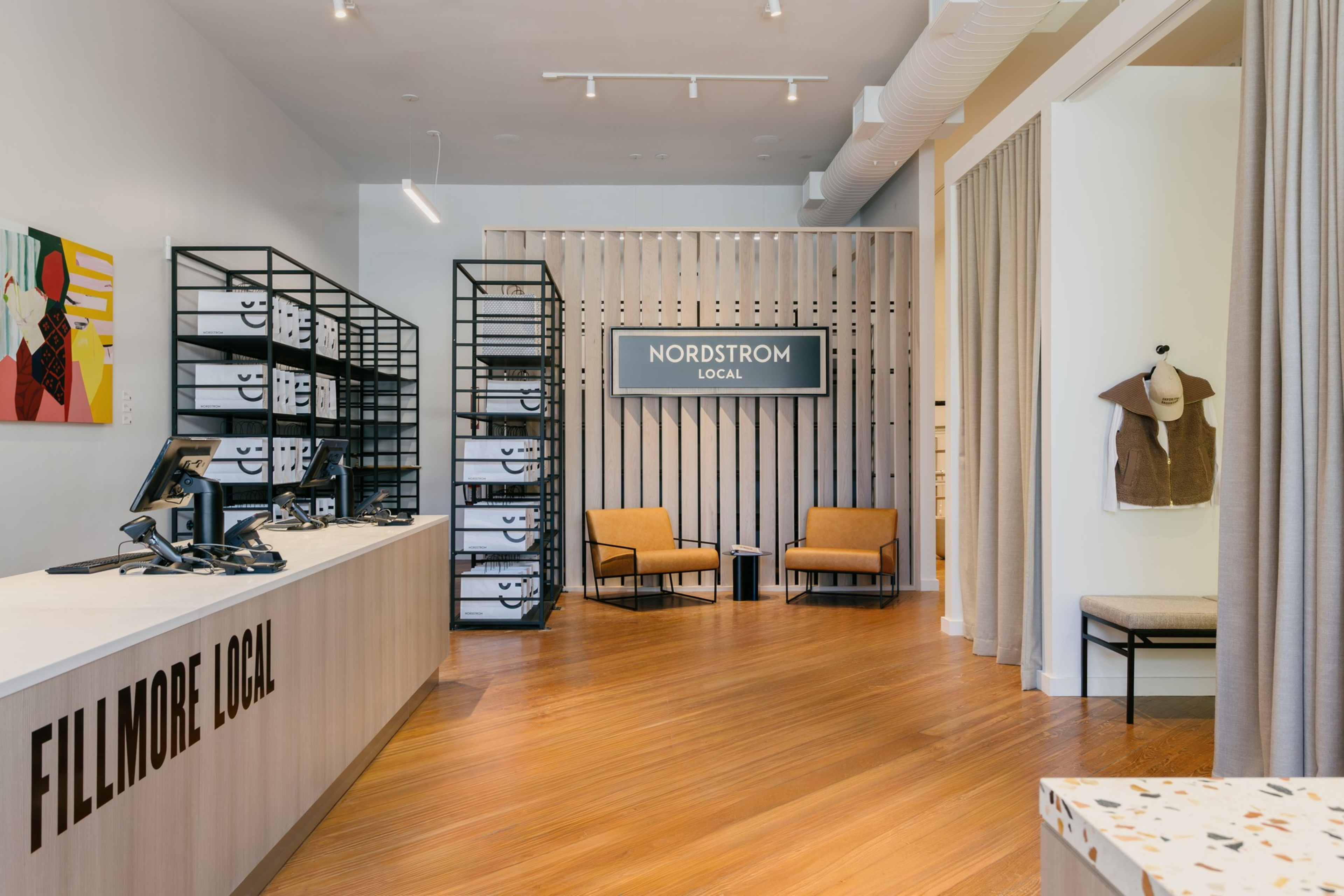 A modern retail space with wooden floors, a “FILLMORE LOCAL” counter, black metal shelves holding bags, two orange chairs, and a “NORDSTROM LOCAL” sign.